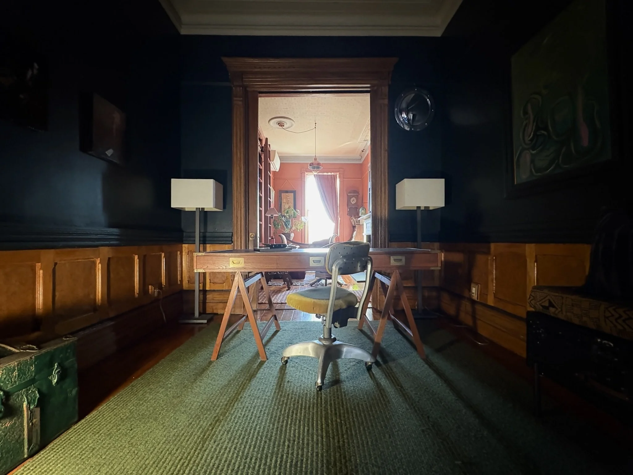 A vintage-style home office with a wooden desk, an office chair, two white lamps on either side of the desk, a green carpet, and a view into a sunlit living room.