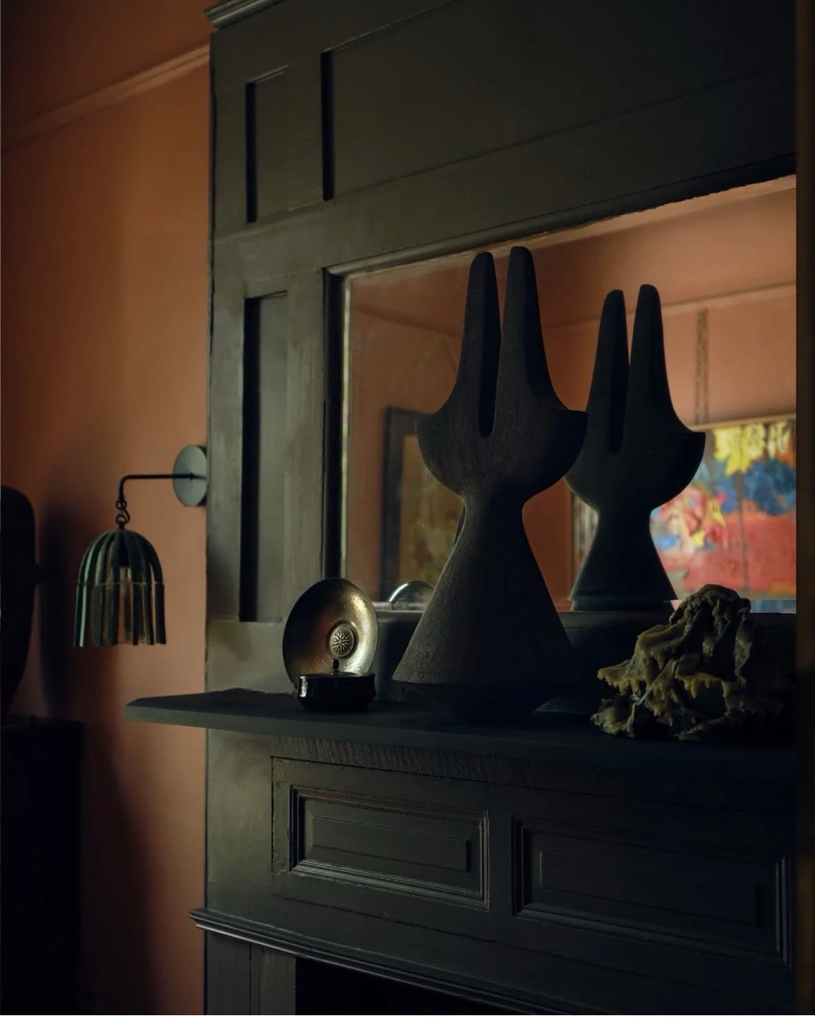 Decorative wooden sculpture resembling abstract animal figures on a dark-colored sideboard, with a small metallic ornament and a shell-like object beside it, reflected in a mirror, in a room with orange walls and a colorful painting in the background