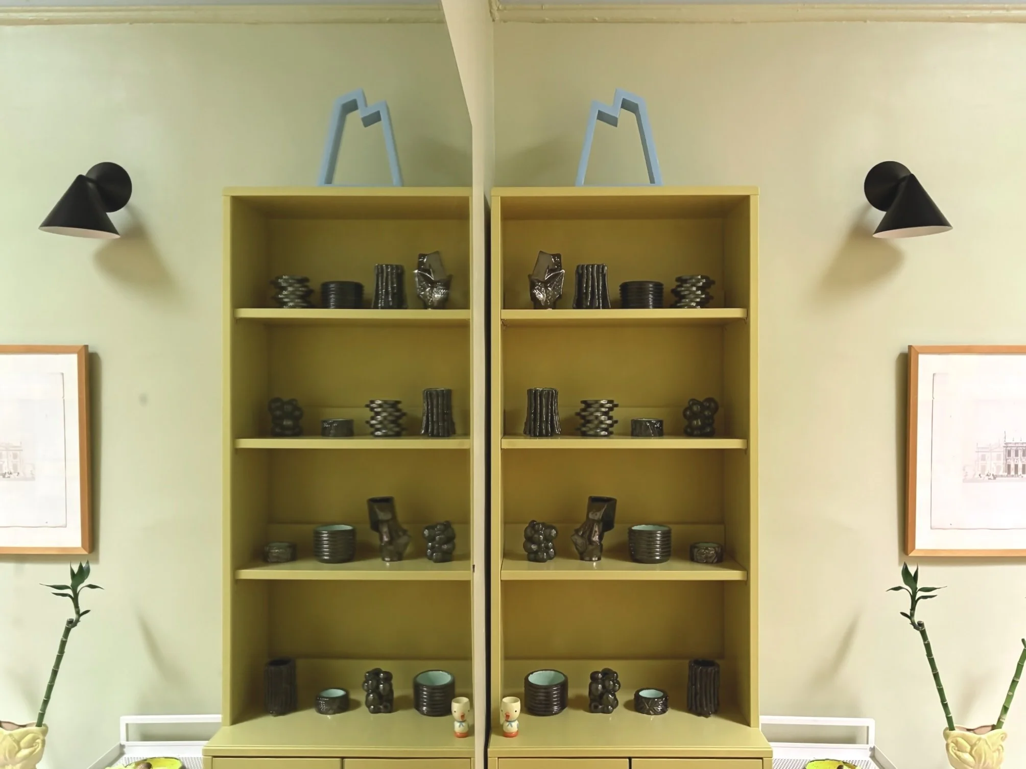 Two yellow bookshelves filled with black and brown ceramic pottery, positioned against a light green wall with framed artwork and black wall-mounted lamps on either side.