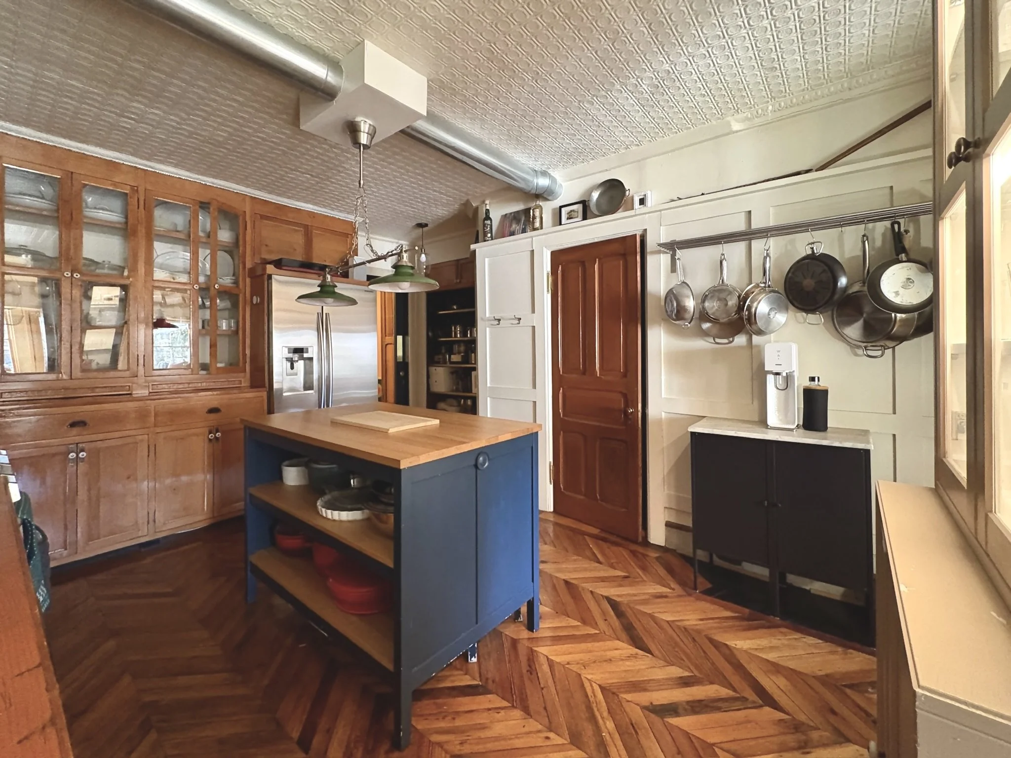 A cozy kitchen with wooden cabinets, a central blue kitchen island, a stainless steel refrigerator, and a hanging pot rack with various pots and pans. The floor is herringbone hardwood, and the ceiling has decorative tin tiles.