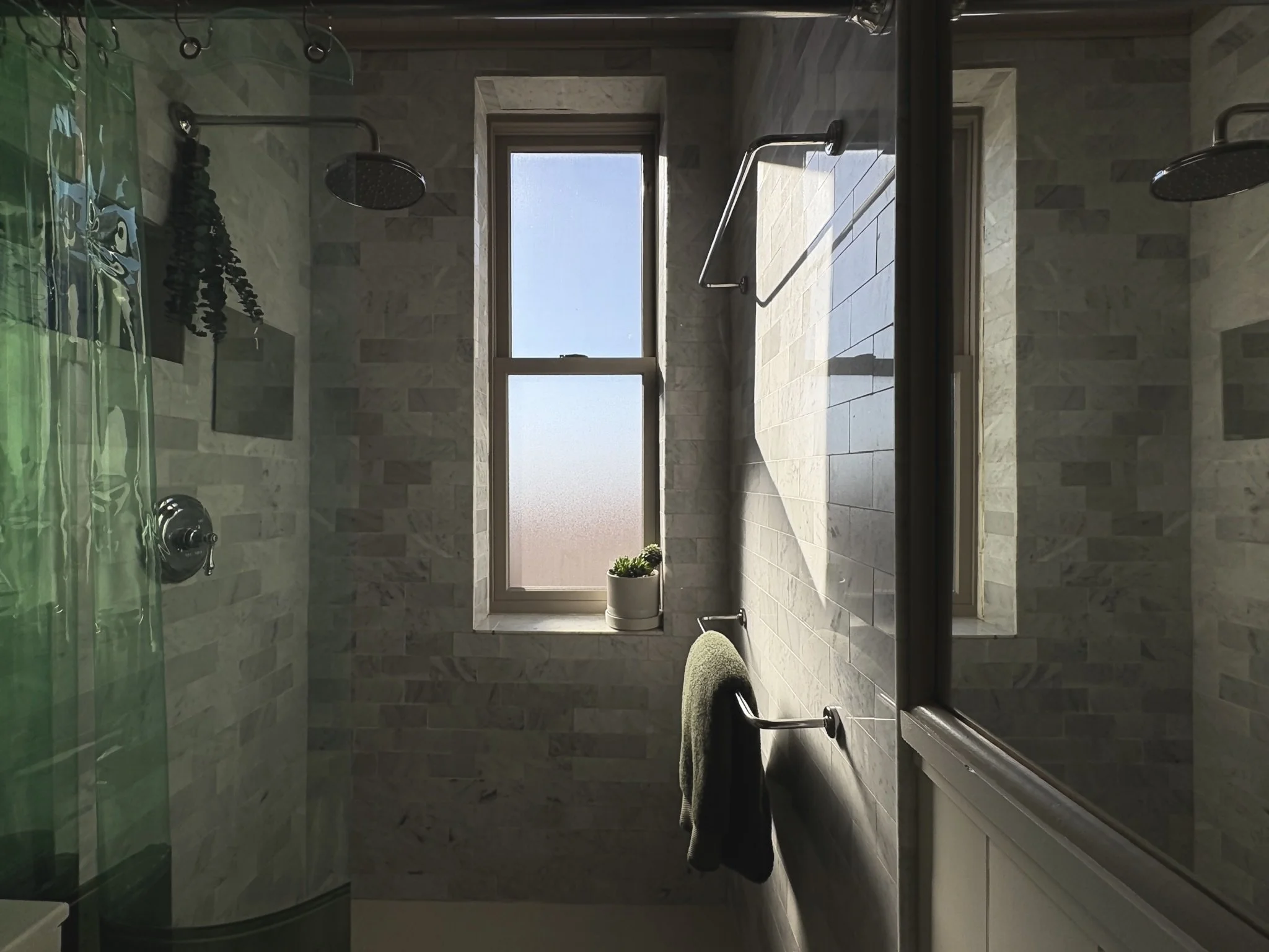 Bright bathroom with two windows, a potted plant, towel rack with a green towel, showerhead, and a glass shower enclosure with a green shower curtain.