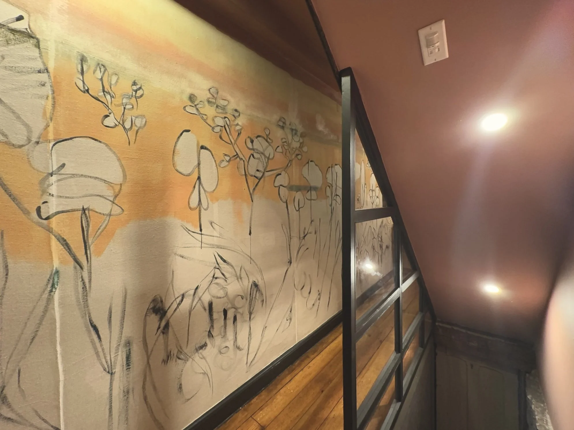 Interior of a staircase with a mural painting of abstract floral designs and a metal railing, illuminated by ceiling lights.