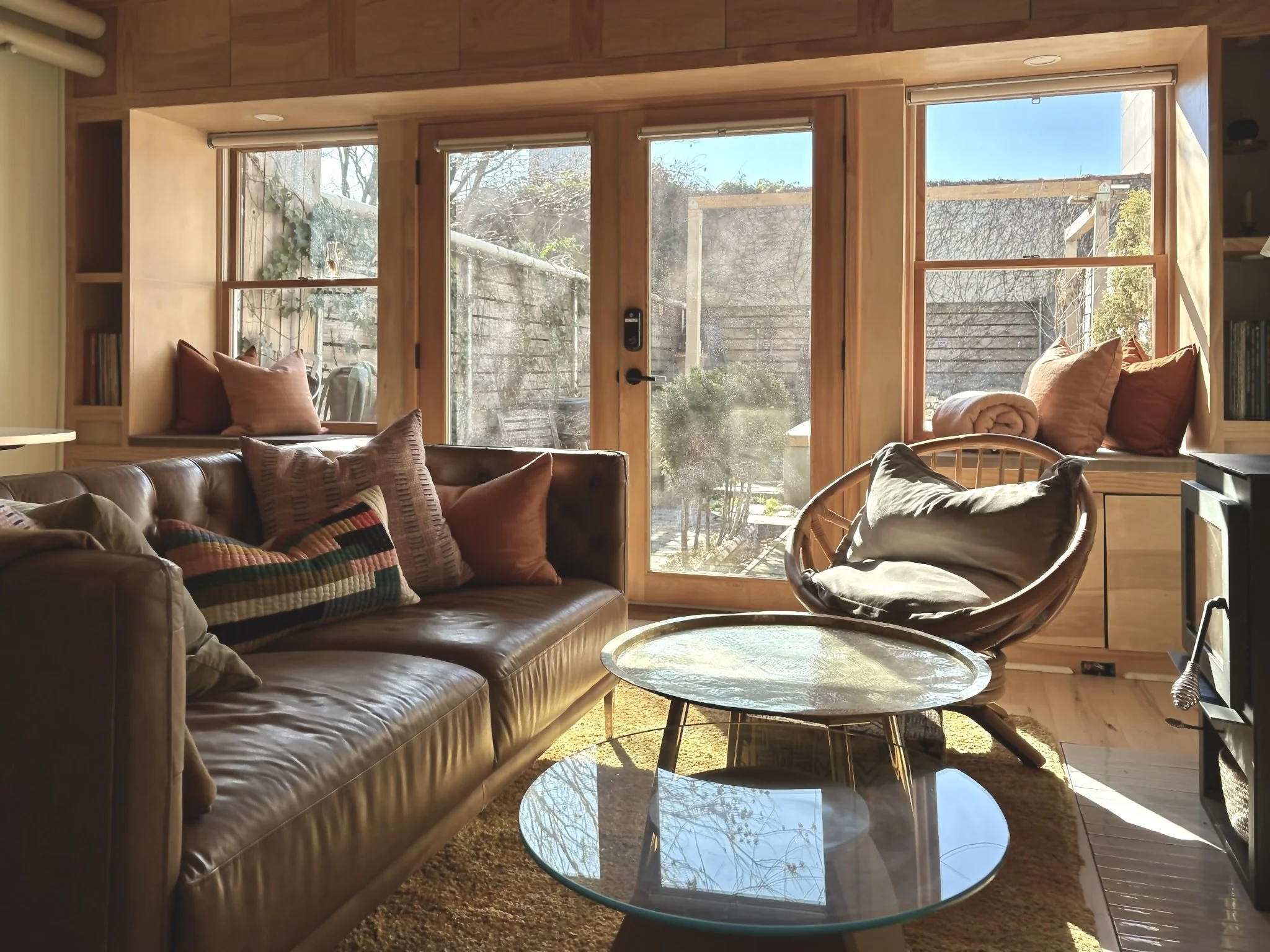 Living room with large window seat and a view of a backyard with trees, featuring a leather sofa with pillows, a modern lounge chair with a pillow, a glass coffee table, and built-in wooden window seating surrounded by wooden paneling.