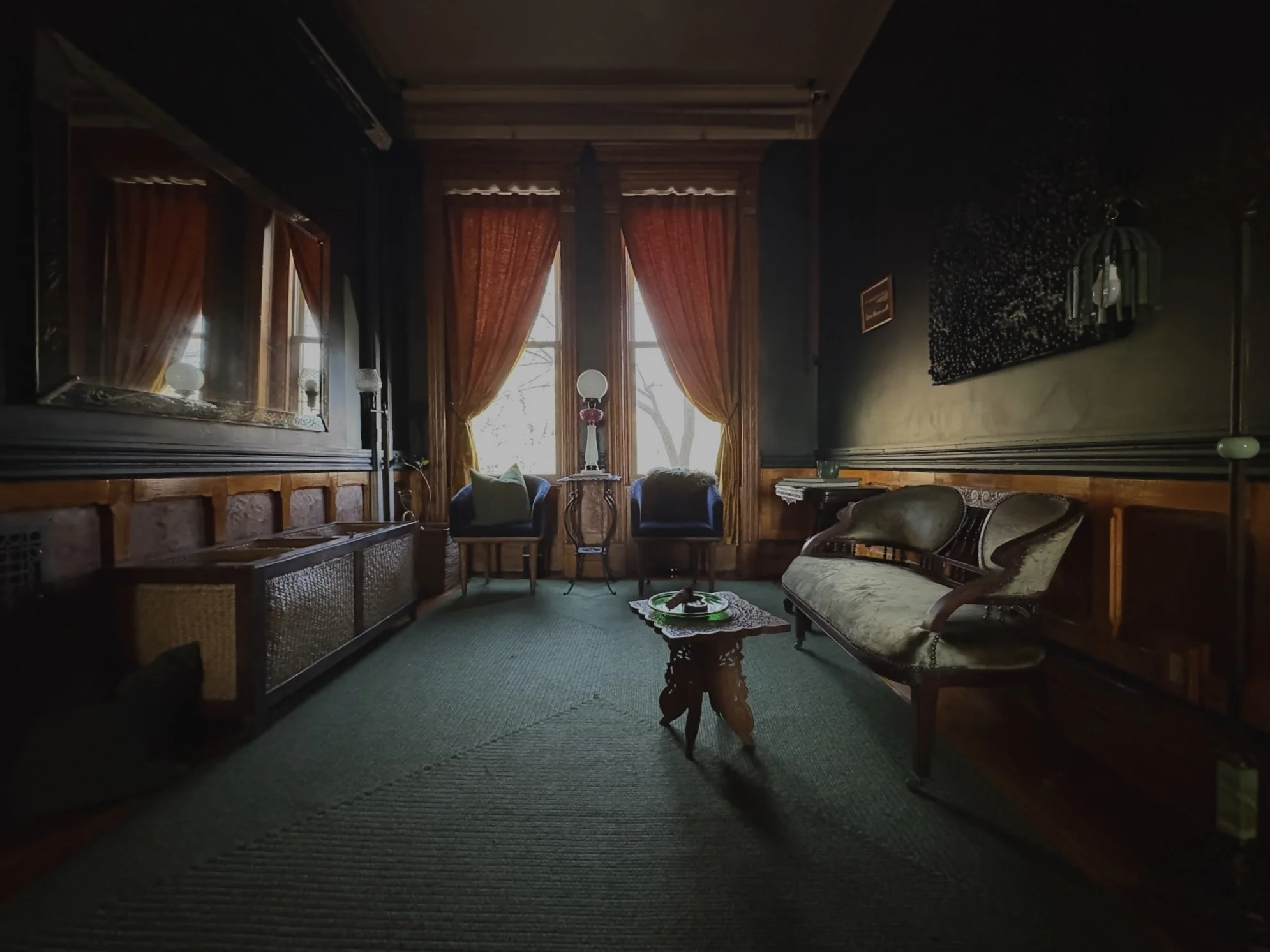 A vintage living room with dark walls, large windows with orange curtains, a green carpet, an ornate sofa, armchairs, a small wooden table, and decorative lamps.