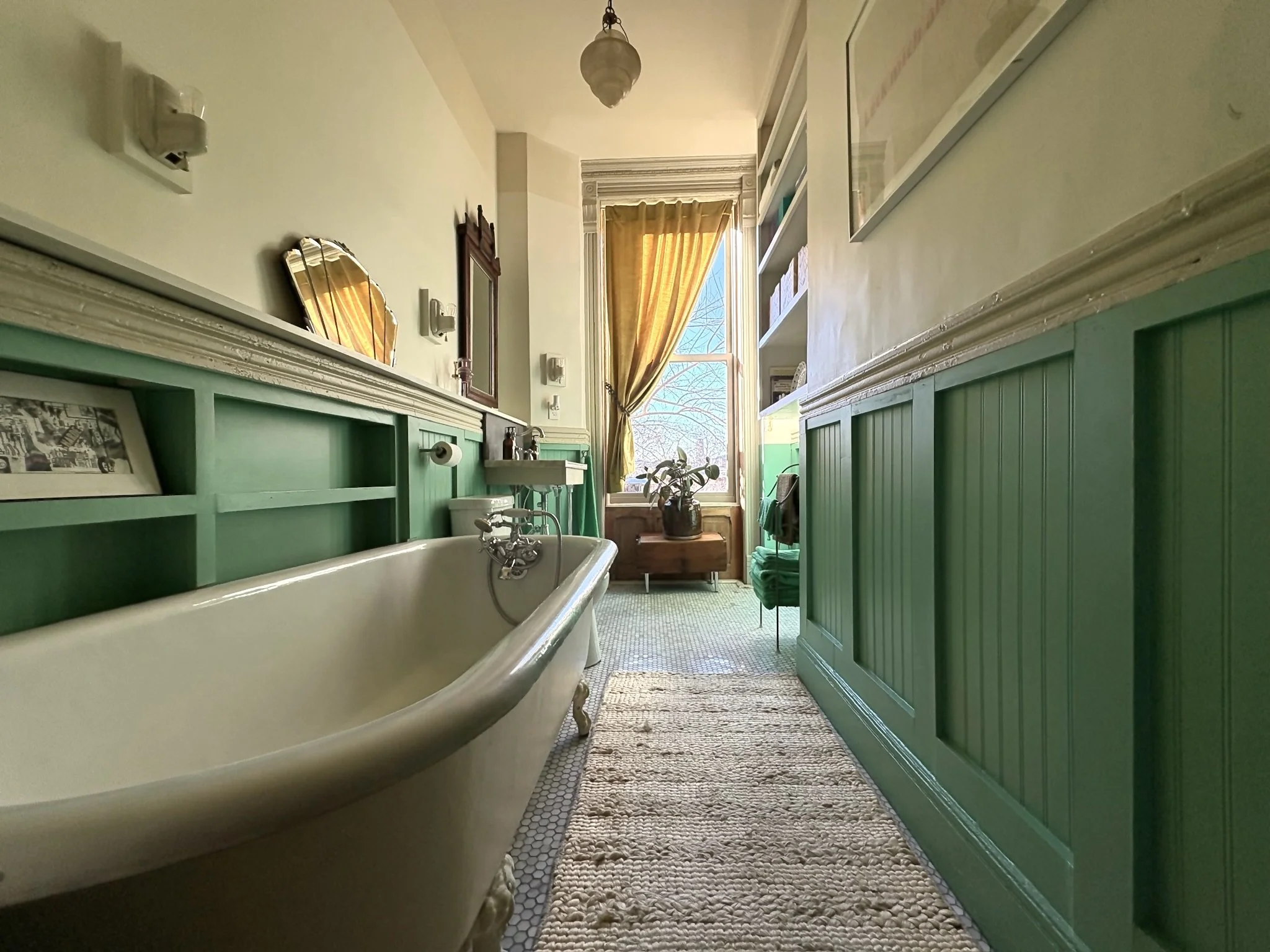 A vintage-style bathroom with a clawfoot bathtub on the left, white walls with green wainscoting, a window with yellow curtains, a potted plant, and a beige rug on the floor.