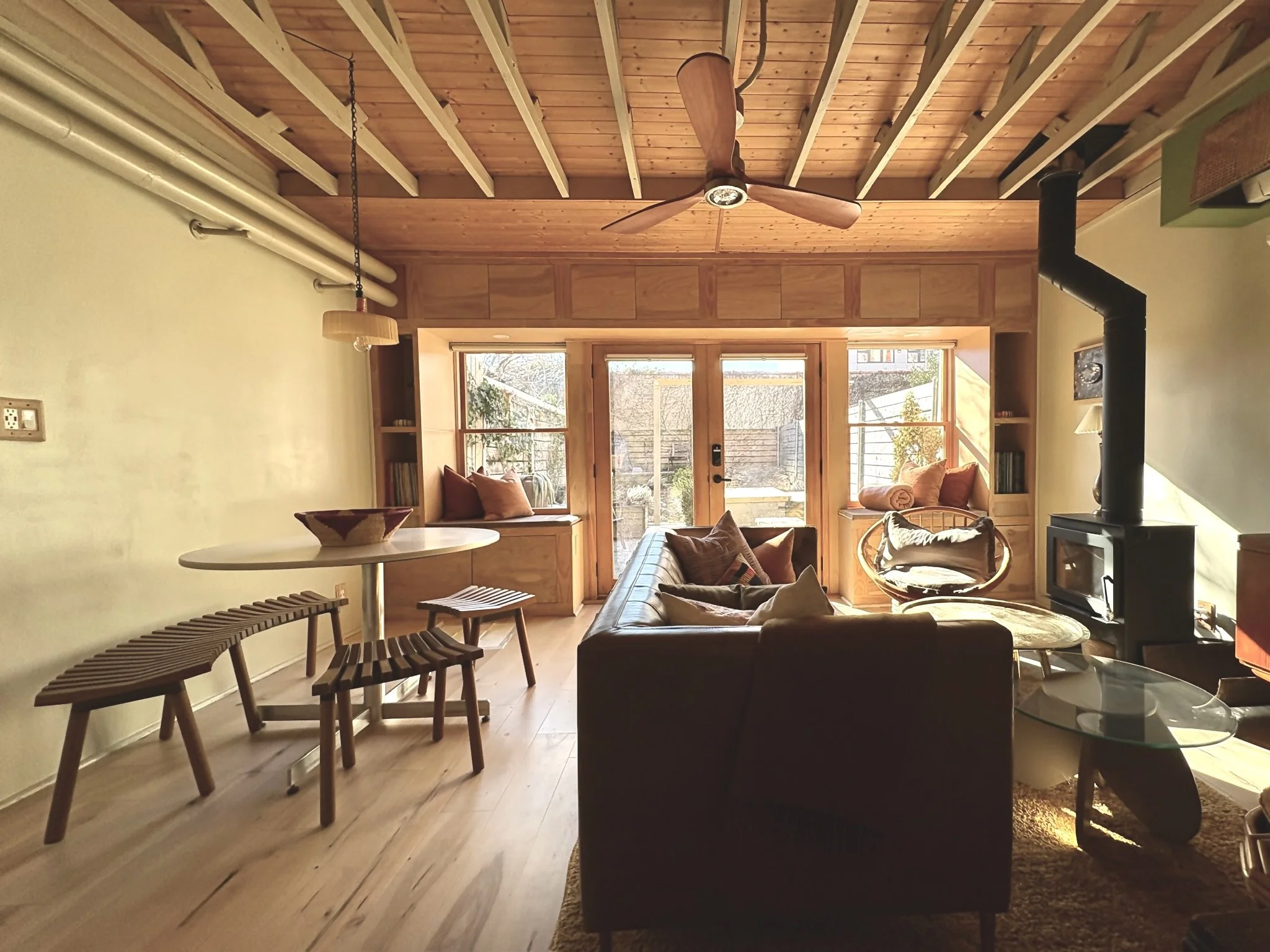 A cozy living room with wooden accents, a large window seat with cushions, a leather sofa, a round glass coffee table, and a wood stove against the right wall, illuminated by warm natural sunlight.