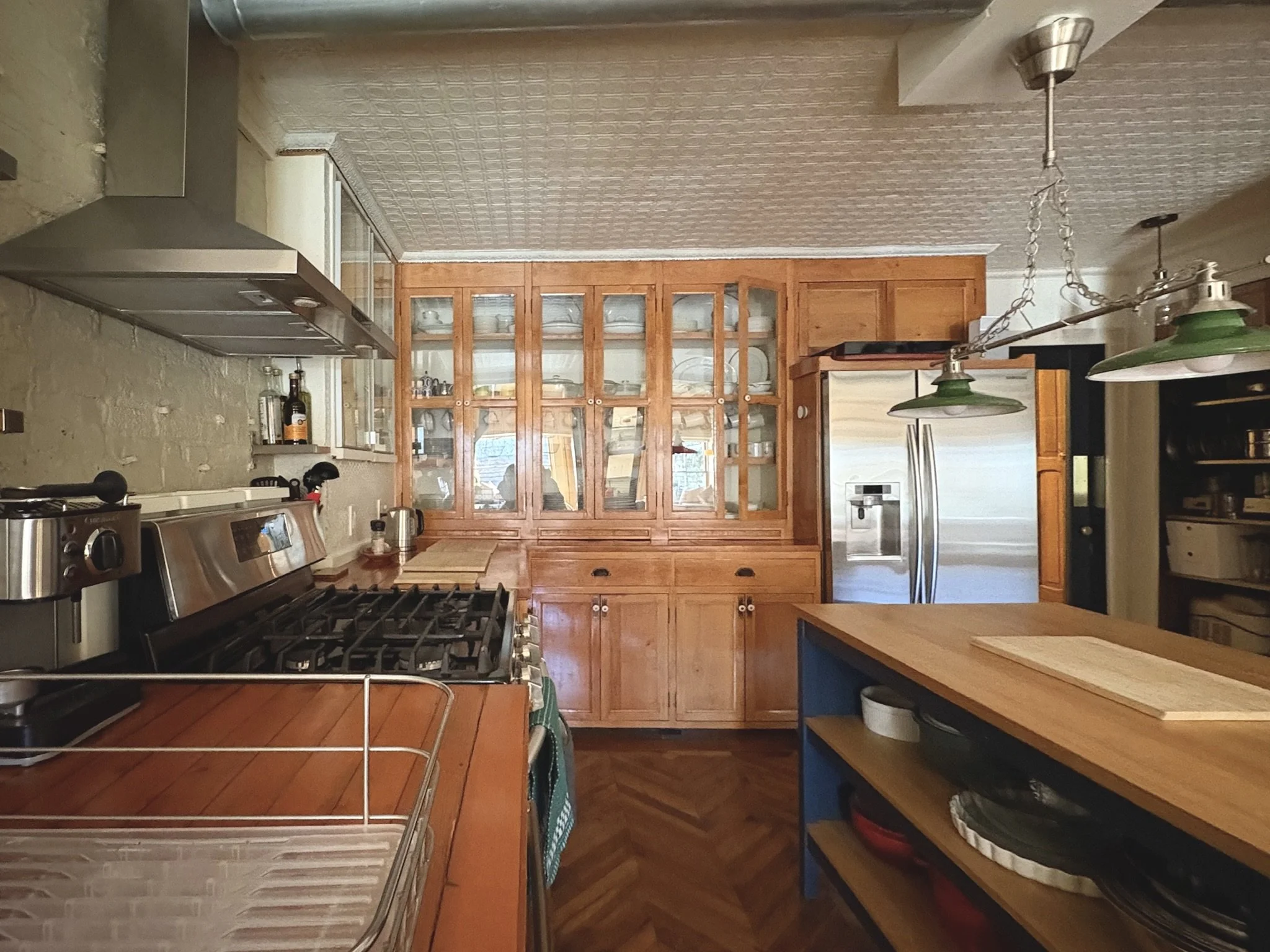 Kitchen with wooden cabinets, stainless steel appliances, and a wooden island, featuring a herringbone-patterned wooden floor and hanging green pendant lights.