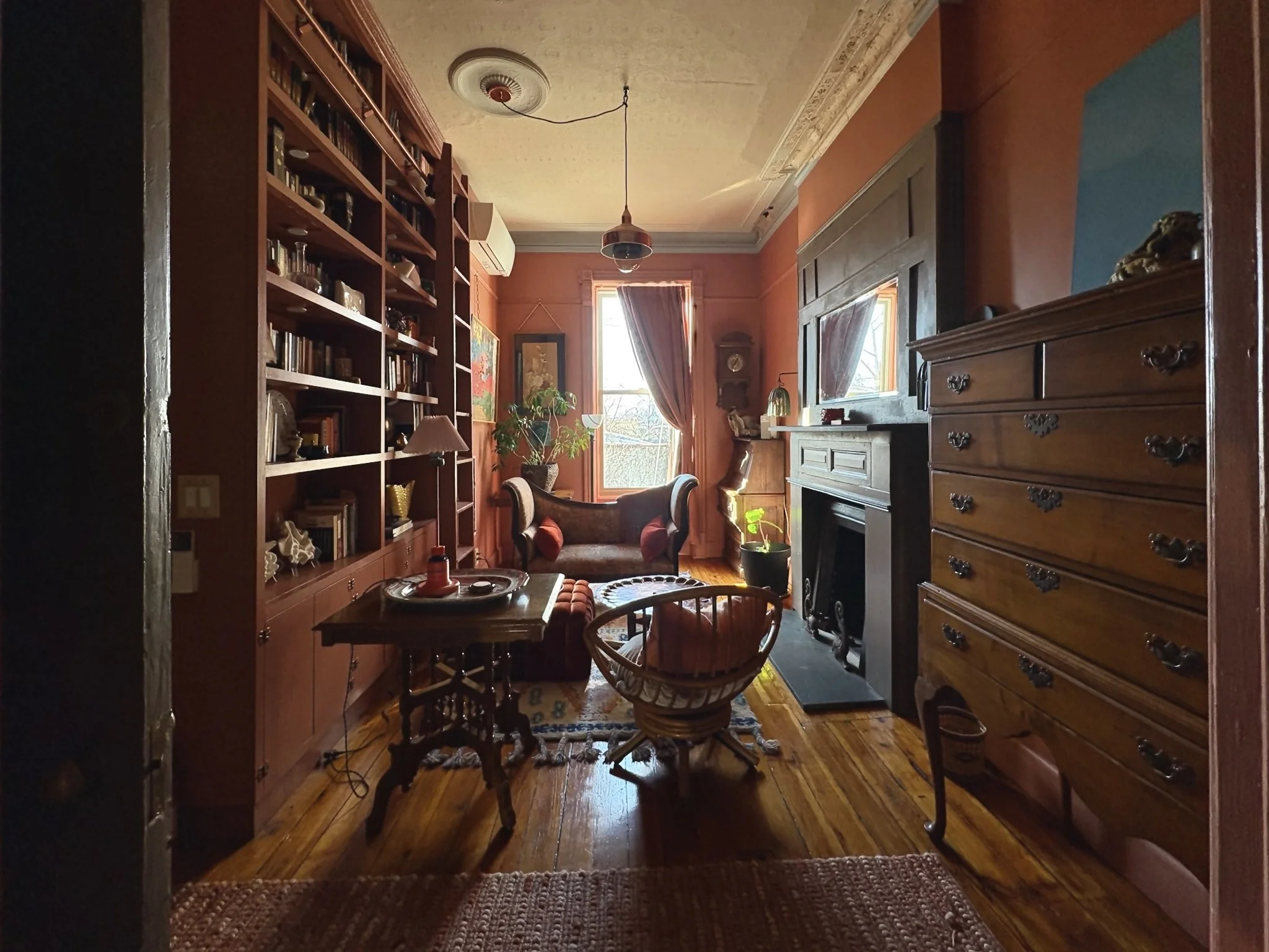 A cozy living room with warm orange walls, wooden furniture, bookshelves, and a fireplace, illuminated by natural light from windows, decorated with plants and artwork.