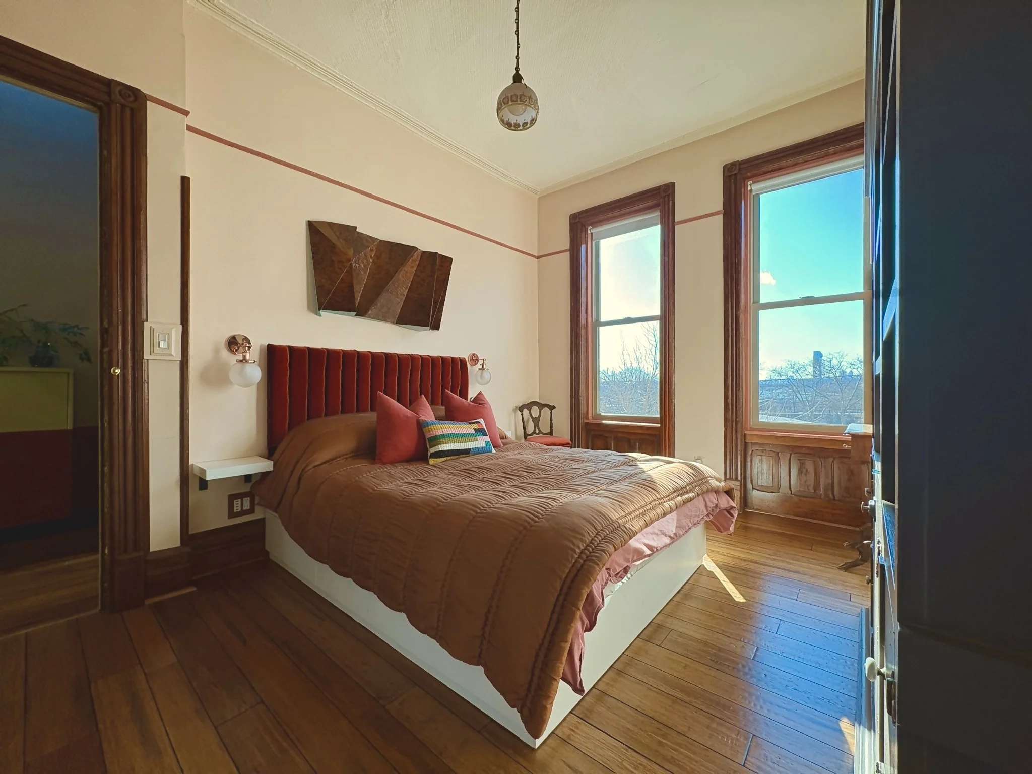 Cozy bedroom with wooden floors, a red upholstered headboard, a brown bedspread, and colorful pillows. Large windows let in natural light, with trees visible outside. Wall-mounted lamps and a modern artwork above the bed add decor.