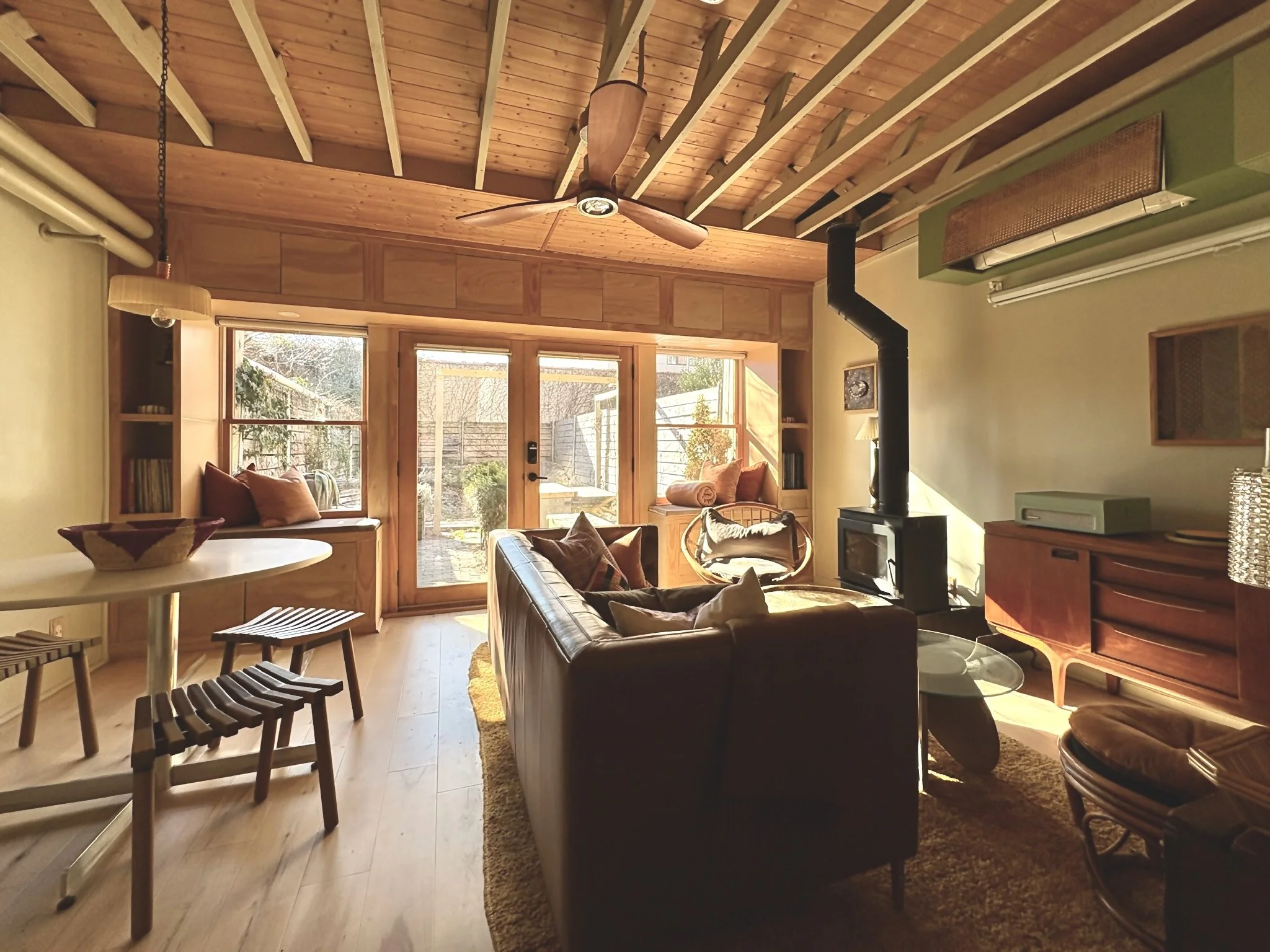 Living room with wood-paneled ceiling, large windows, and a glass door leading outside. Contains a leather sofa, wooden sideboard, small round table, chairs, a wood stove, and built-in bookcases. Sunlight filters in, creating a warm atmosphere.