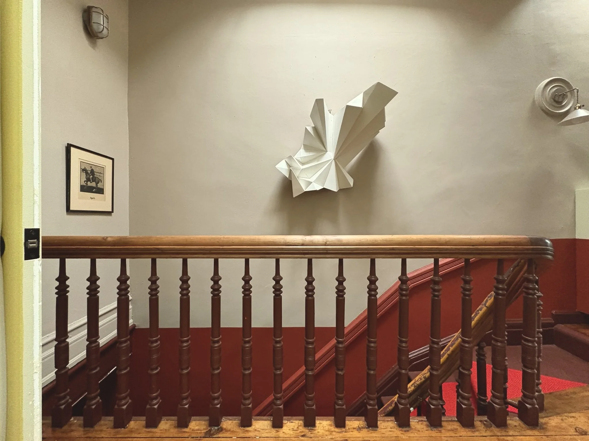 Interior of a stairwell with a wooden handrail, a framed picture on the wall, a white modern art sculpture resembling a butterfly or geometric figure, and wall-mounted lights. The walls are painted in light and dark tones, and red carpet runs along t