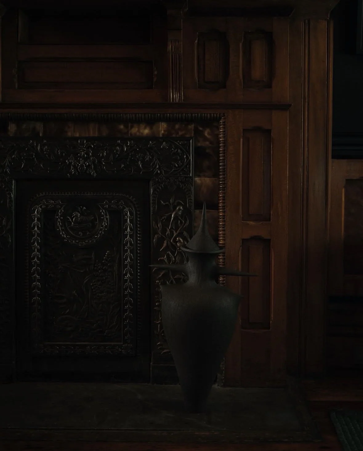 Dark intricately carved fireplace surround with floral motifs, next to a tall, dark, curved vase with a spire-top, set against a rich wood-paneled wall.