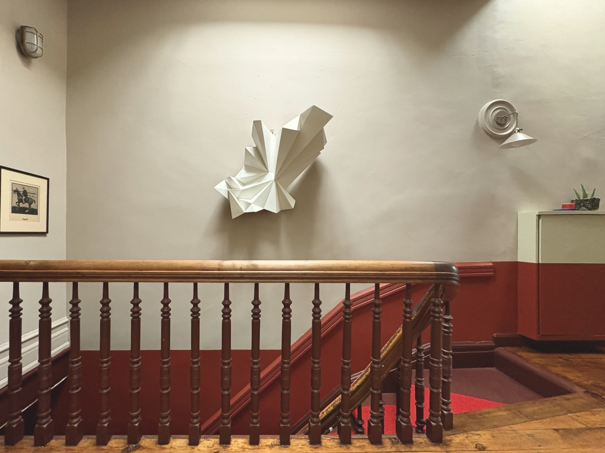 An interior staircase with a wooden handrail and red carpet. The wall behind features a middle-sized sculpture of a butterfly or moth with abstract, geometric wings, and a small framed black and white picture of a person on horseback. There is a powe