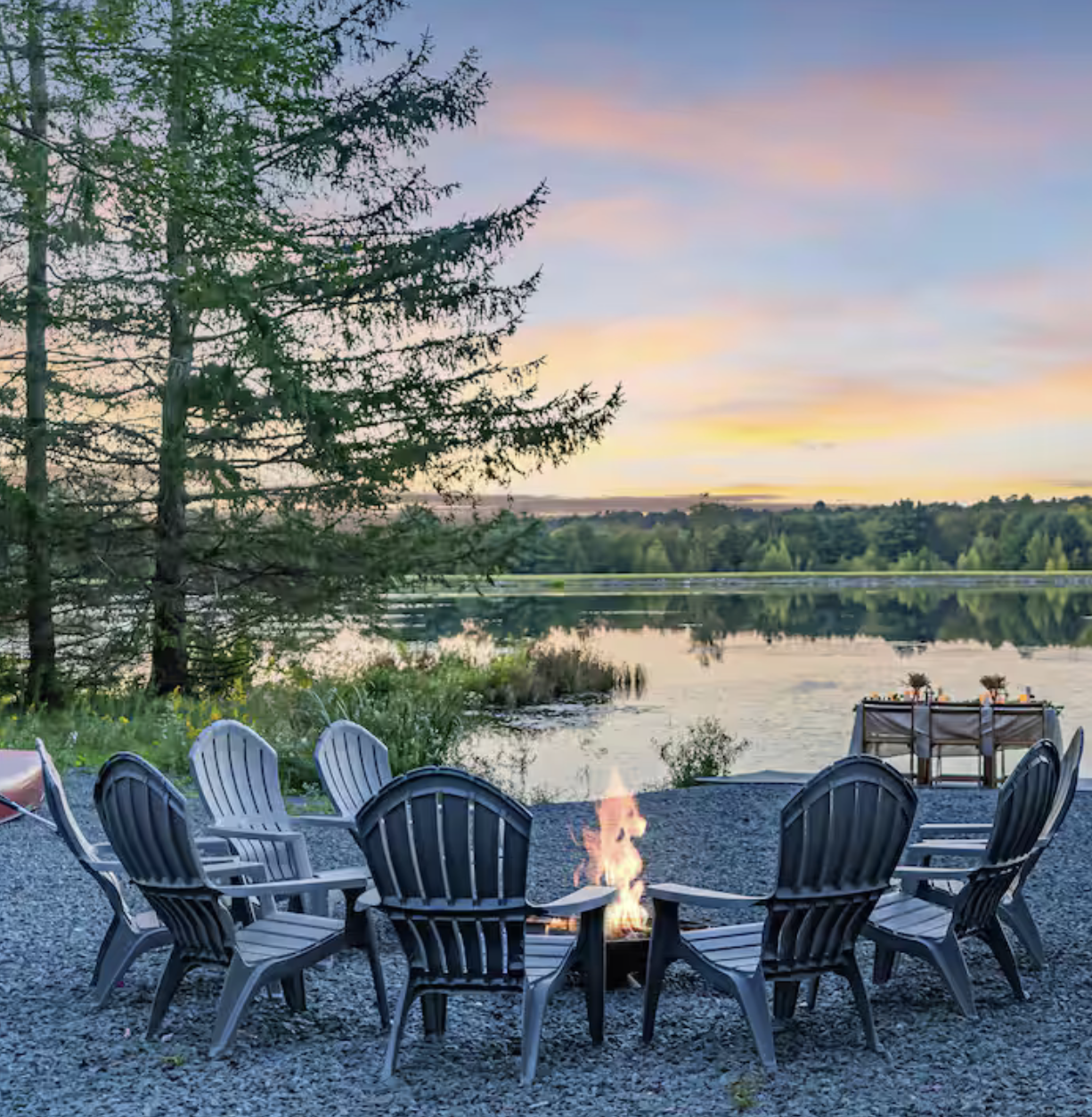 Eight black Adirondack chairs arranged in a circle around a small fire pit with a lake and trees in the background during sunset.