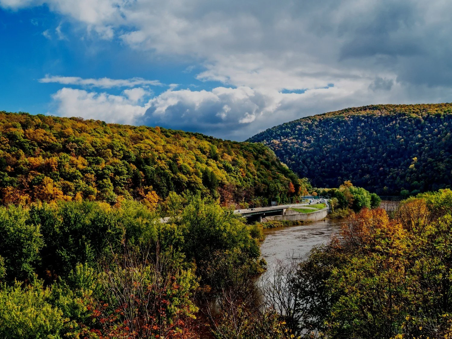 A scenic view of a river flowing through a lush valley surrounded by hills covered in green and autumn-colored trees, with a partly cloudy blue sky overhead.