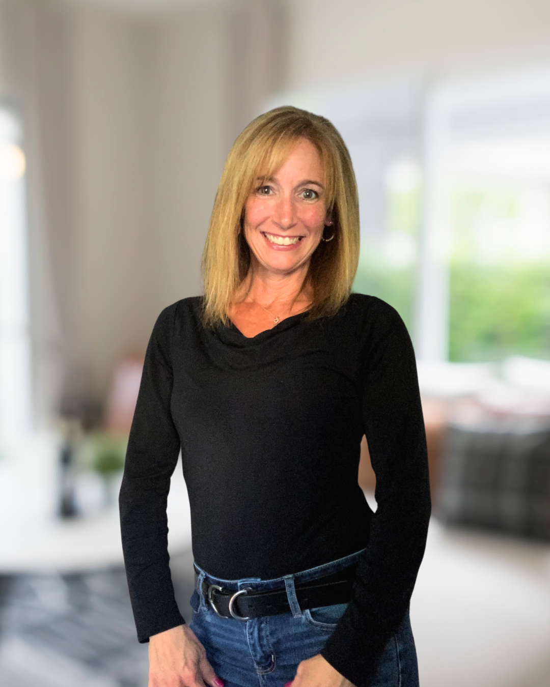 Smiling woman with blonde hair wearing a black top and blue jeans standing in a blurred indoor background.