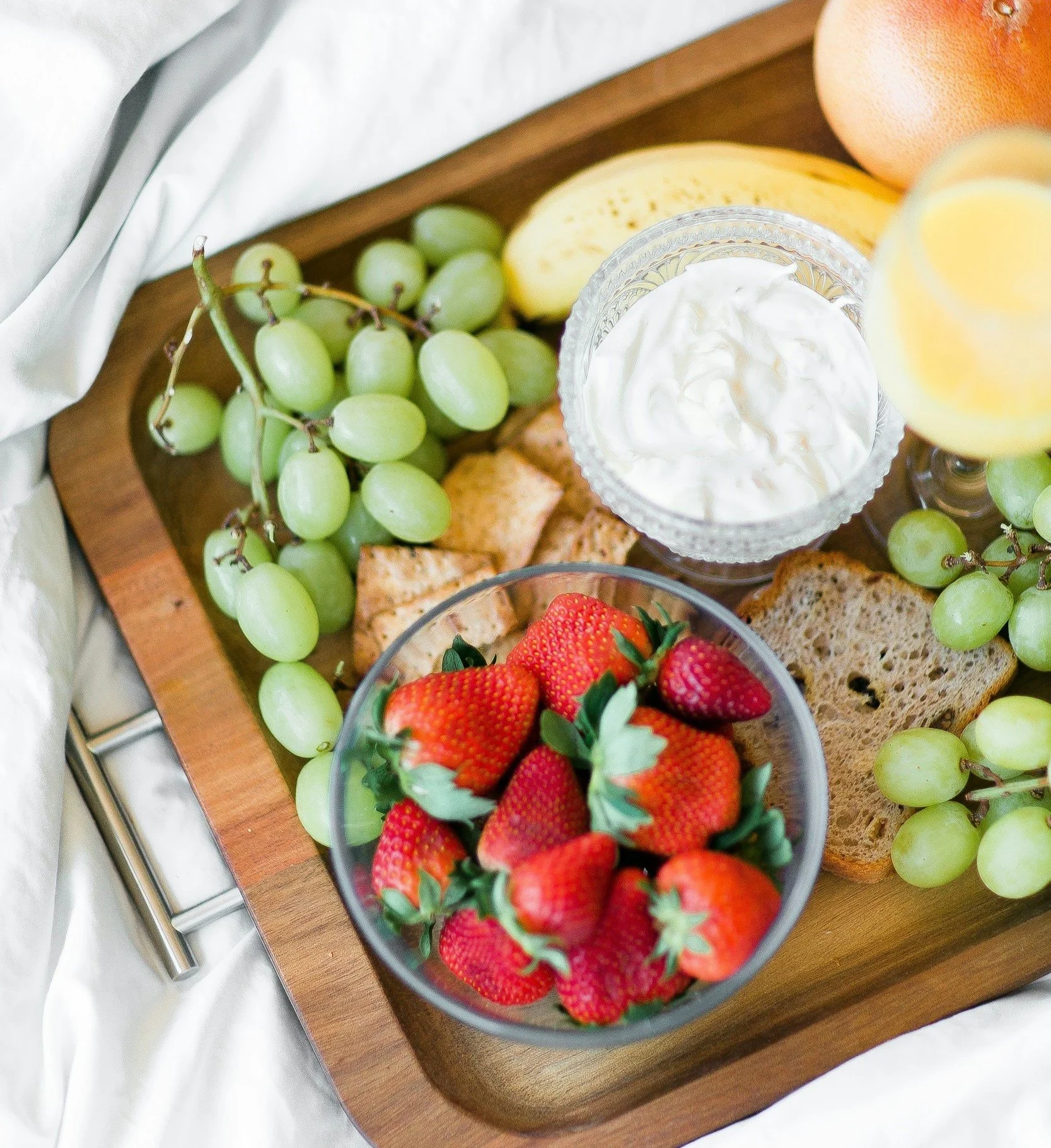 A wooden serving tray with green grapes, strawberries, slices of bread, a banana, a grapefruit, a glass of orange juice, and a bowl of yogurt.