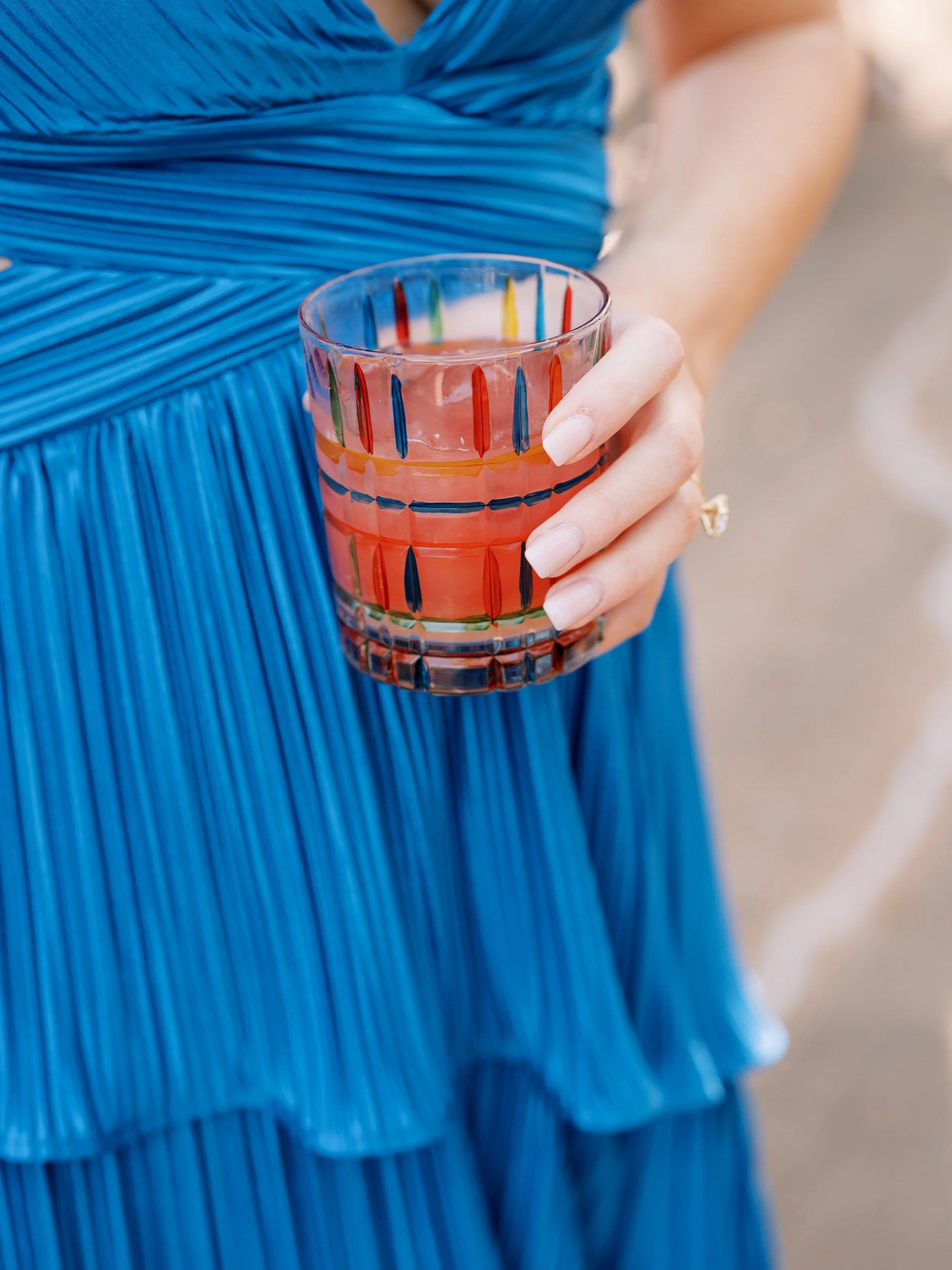 A person wearing a blue pleated dress holding a glass of pink-colored drink with vertical rainbow stripes.