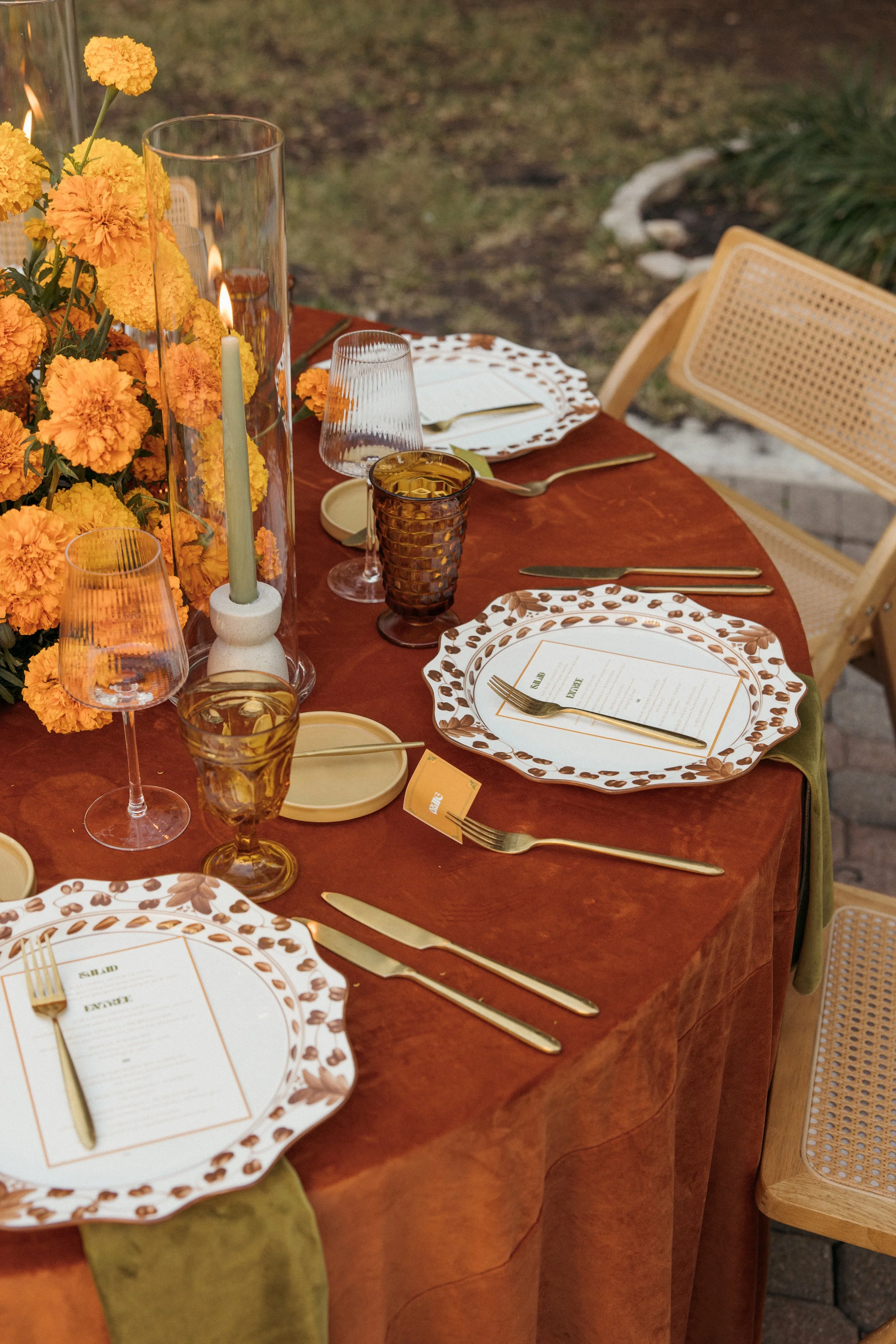A dining table set for a meal with floral plates, gold utensils, wine glasses, and tint glasses, with a centerpiece of yellow flowers and candles, outdoor setting with grass and garden in the background.
