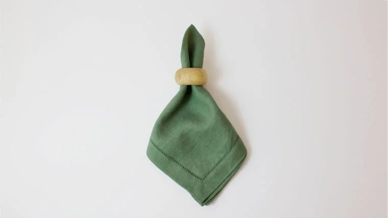 Green cloth napkin tied with a wooden ring in the shape of a bunny ear on a plain white background.
