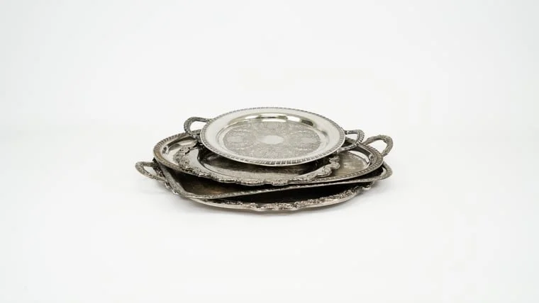 Stack of silver or pewter serving trays with handles on a plain white background.