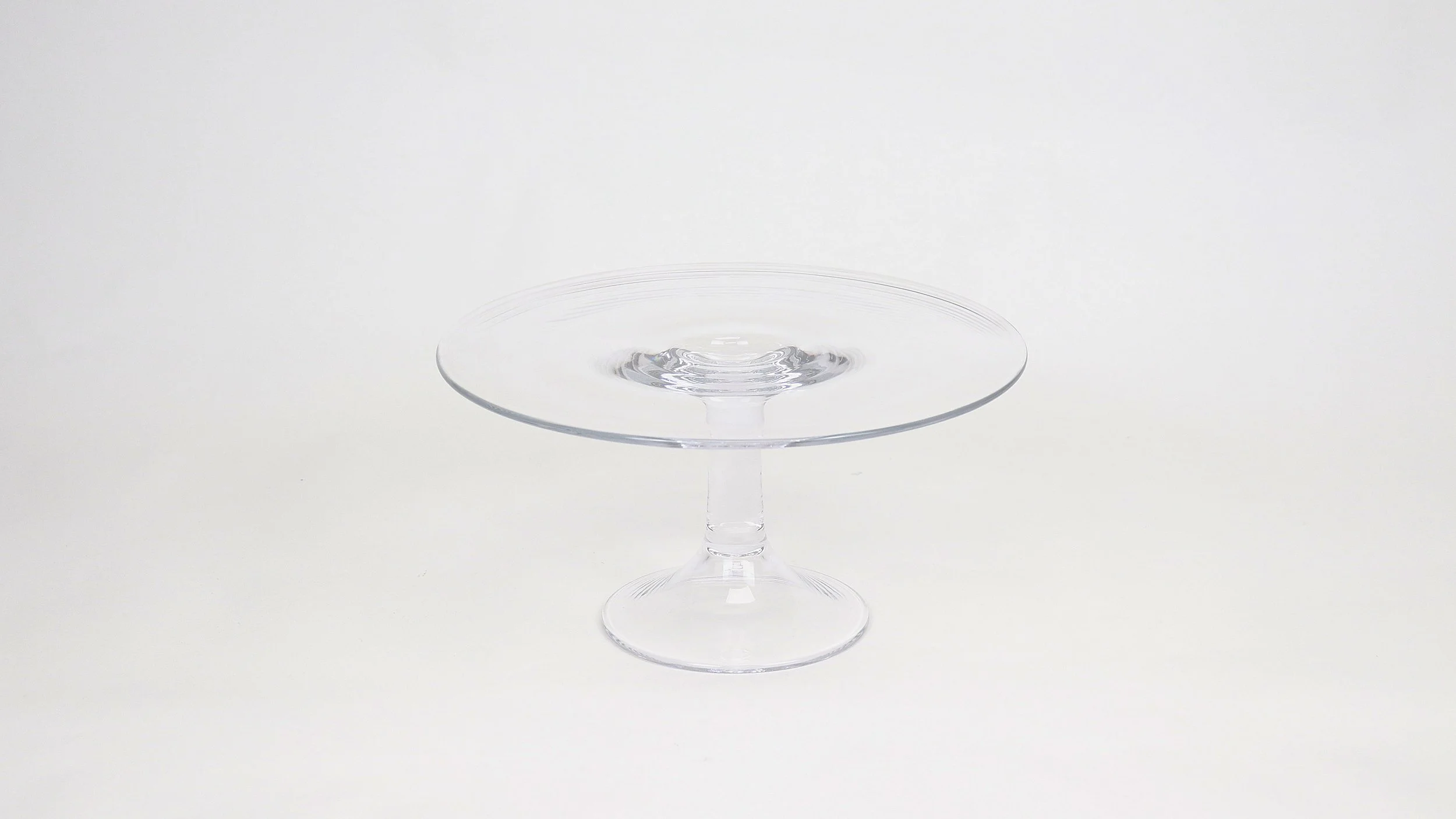 A clear glass dessert stand with a wide flat top and a small base is filled with water, creating a ripple effect on the water surface.
