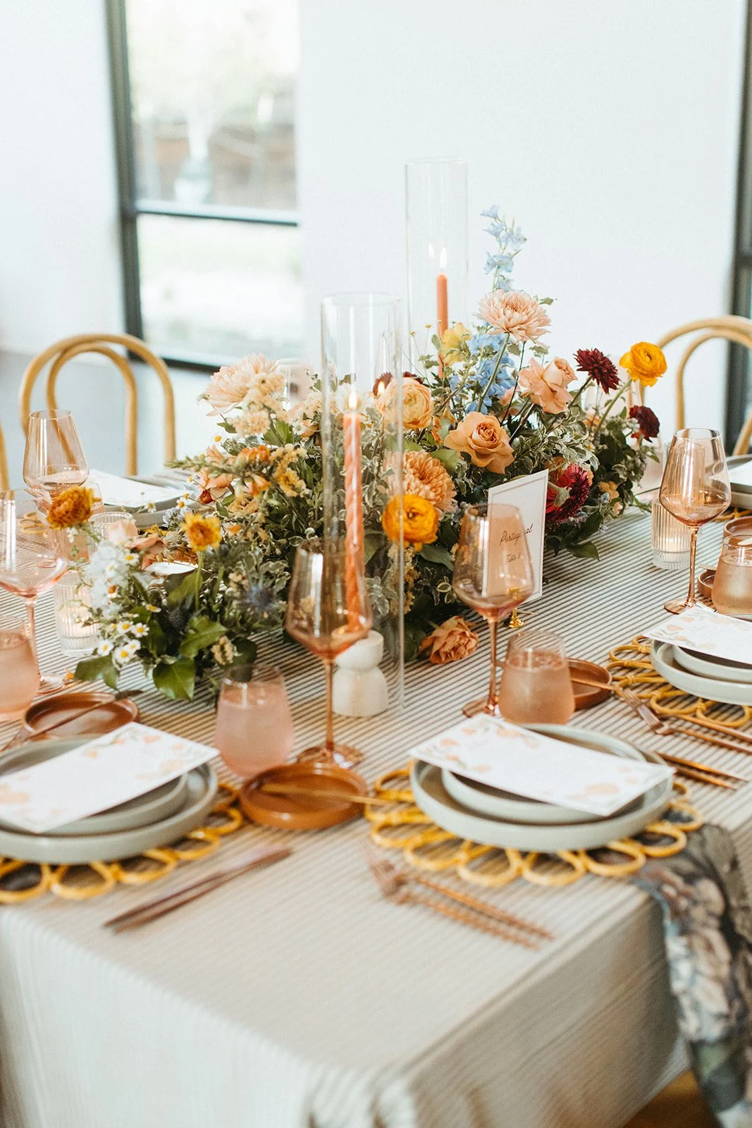 A decorated dining table with pastel-colored plates, pink and rose gold glassware, and a large floral centerpiece with peach, pink, and yellow flowers and greenery.