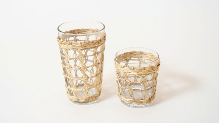 Two clear glass tumblers with woven rattan holders, one taller and one shorter, placed on a white surface.
