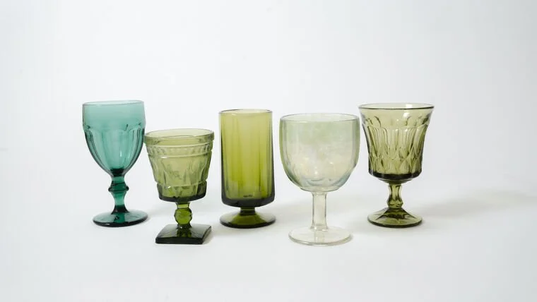 Five vintage glass goblets in shades of green and blue, lined up on a white surface against a plain white background.