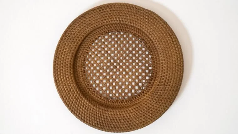 A round, brown, woven rattan tray with a lattice pattern in the center, placed on a white surface.