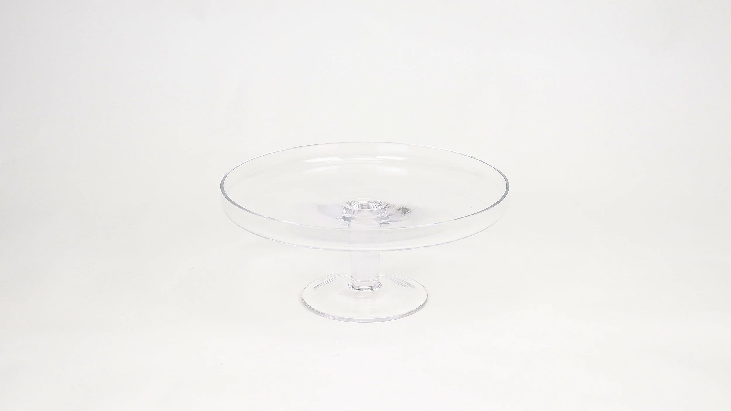 A clear glass dessert stand with a flat, round top and a short stem, set against a plain white background.