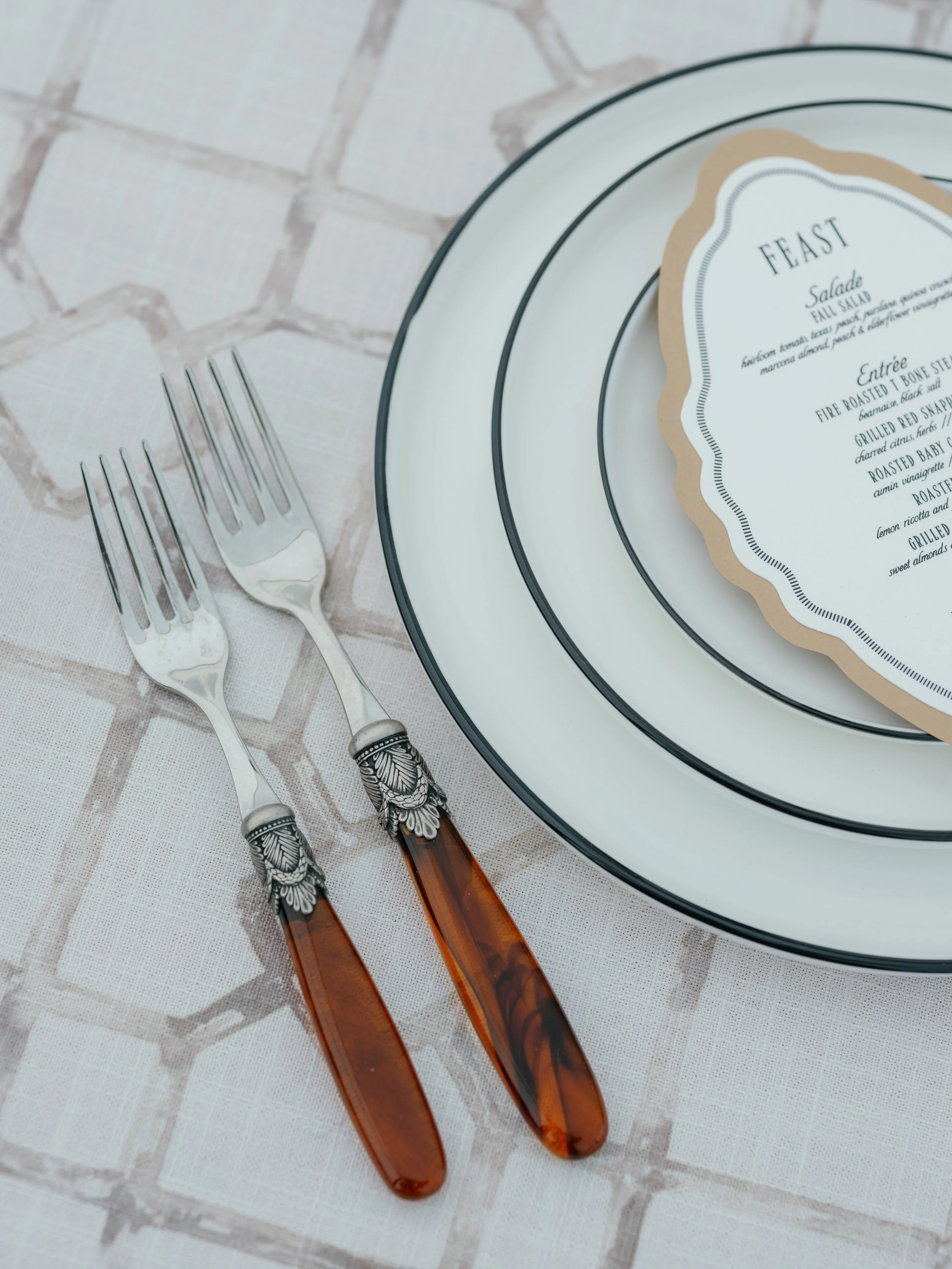 Two forks with decorative handles placed on a tablecloth next to a stack of white plates with black rims and a printed menu in a scalloped beige border.