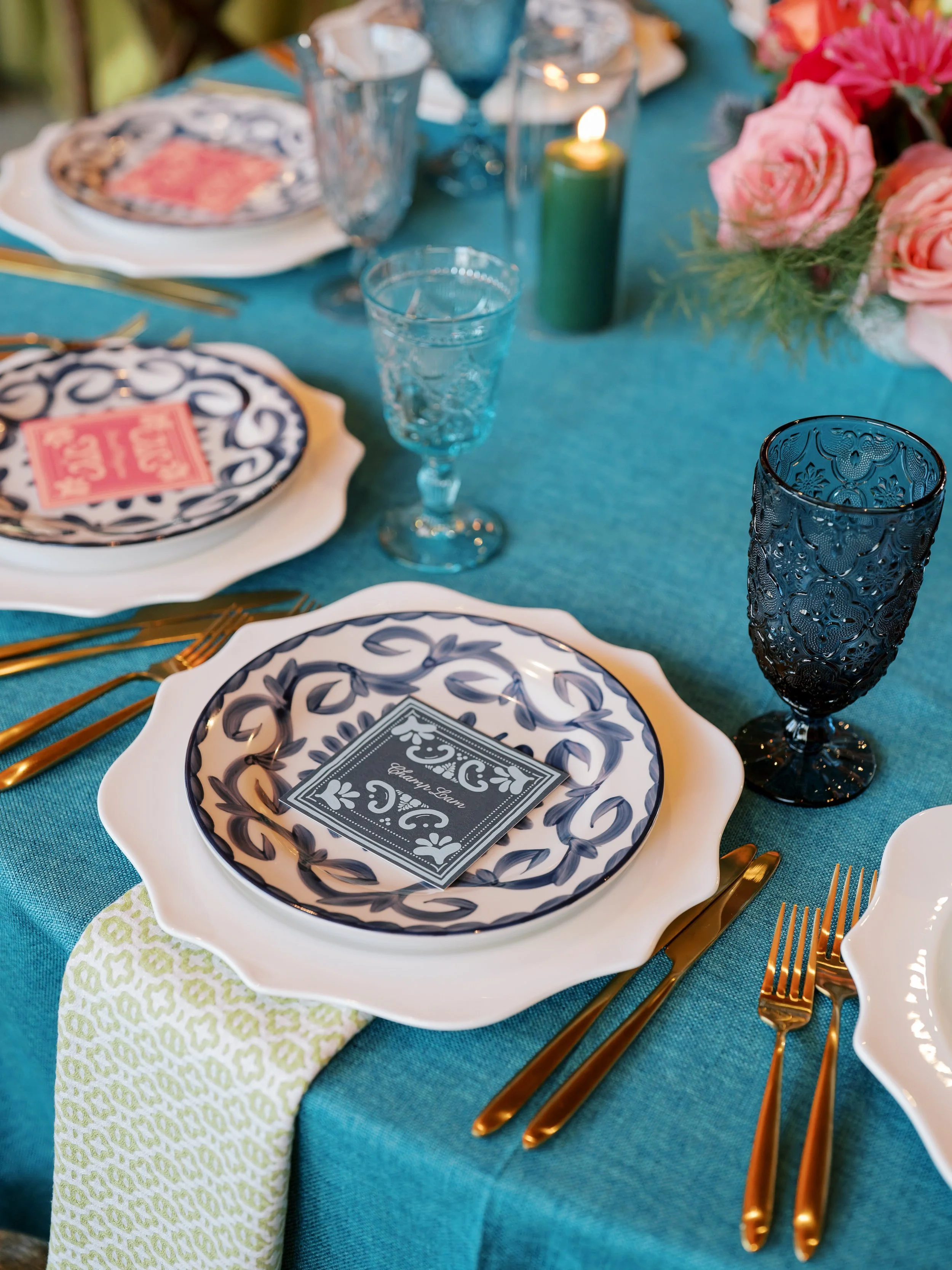 Elegant table setting with patterned plates, gold utensils, blue and black glassware, a green candle, and a bouquet of pink roses on a teal tablecloth.