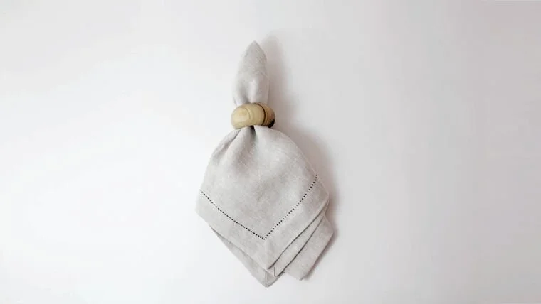 A white cloth napkin tied at the top with a wooden ring, hanging against a plain white wall.