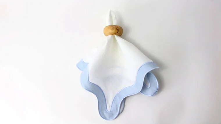 A cloth napkin hanging on a wall with a wooden ring and a small loop on top for hanging, featuring a white main body and blue border.