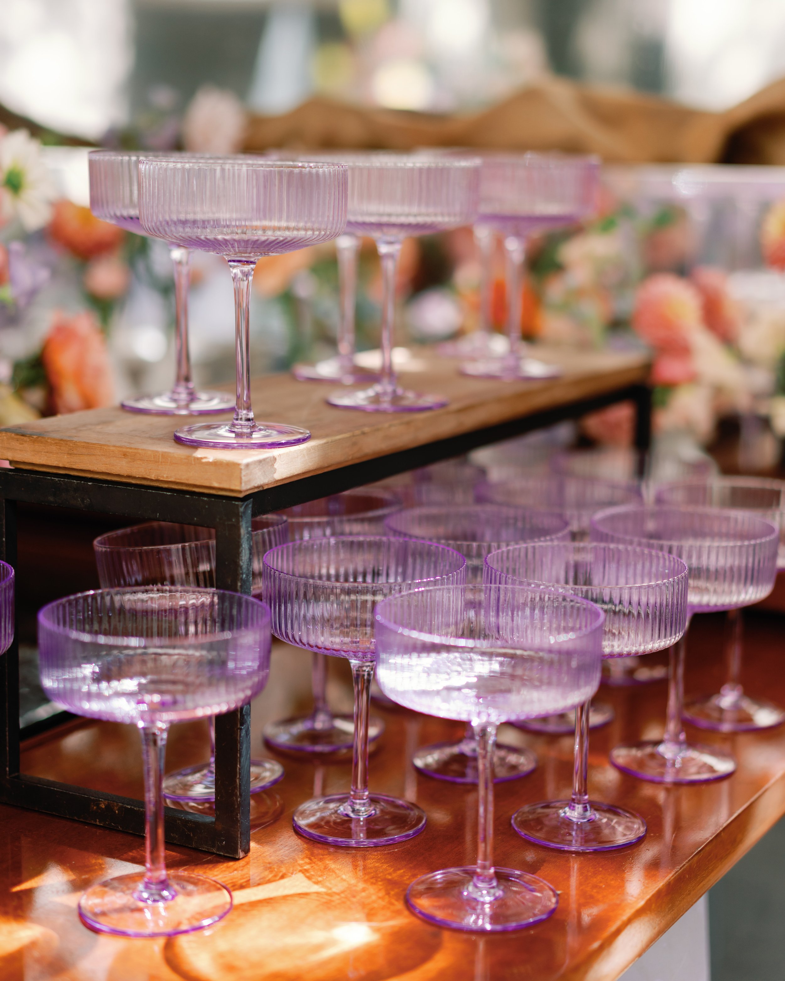 Multiple pink-colored cocktail glasses with ridged design on a wooden surface, some arranged on a black stand, with blurred background featuring flowers and other decorative items.