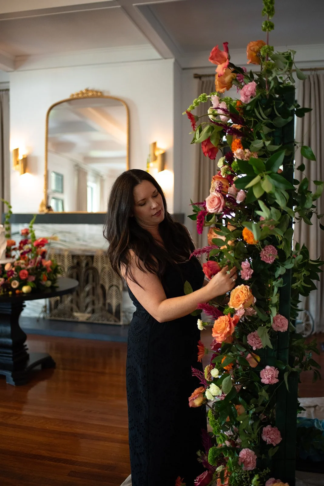 A woman in a black dress arranging pink, orange, and red flowers on a vertical floral display in a well-lit interior room.