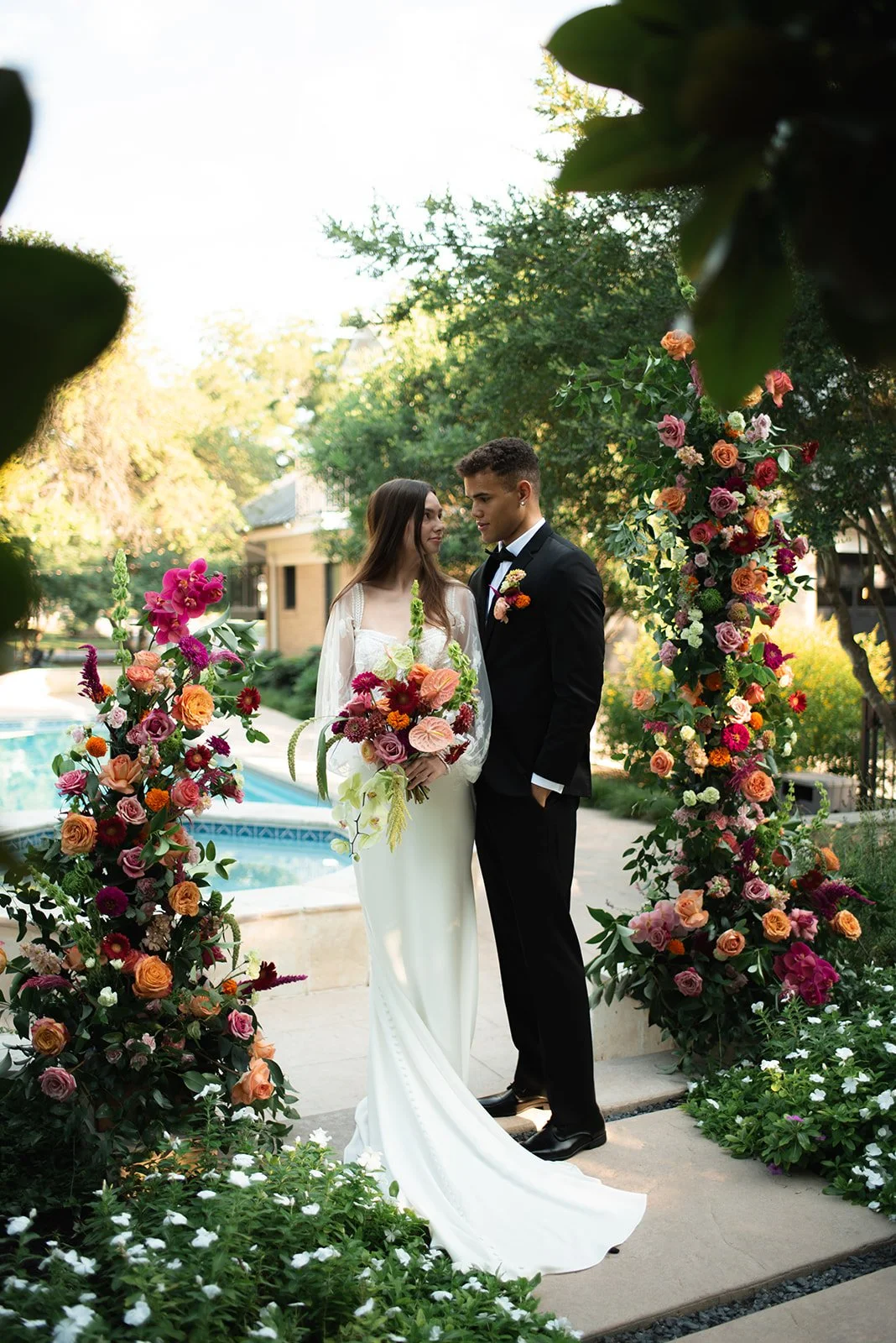 A bride and groom stand together among colorful floral arrangements at an outdoor wedding ceremony, with trees and a pool in the background.