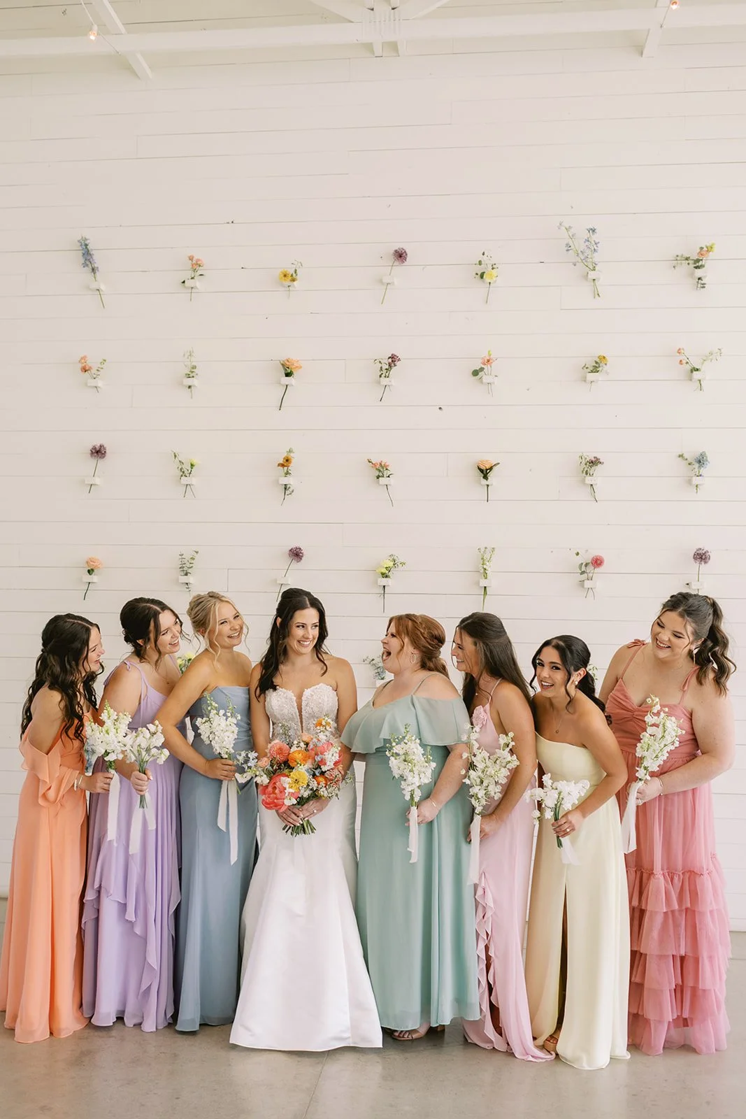 Group of women at a wedding, wearing pastel-colored dresses, standing against a white wall decorated with flower arrangements. The bride is in the center holding a bouquet, surrounded by bridesmaids.