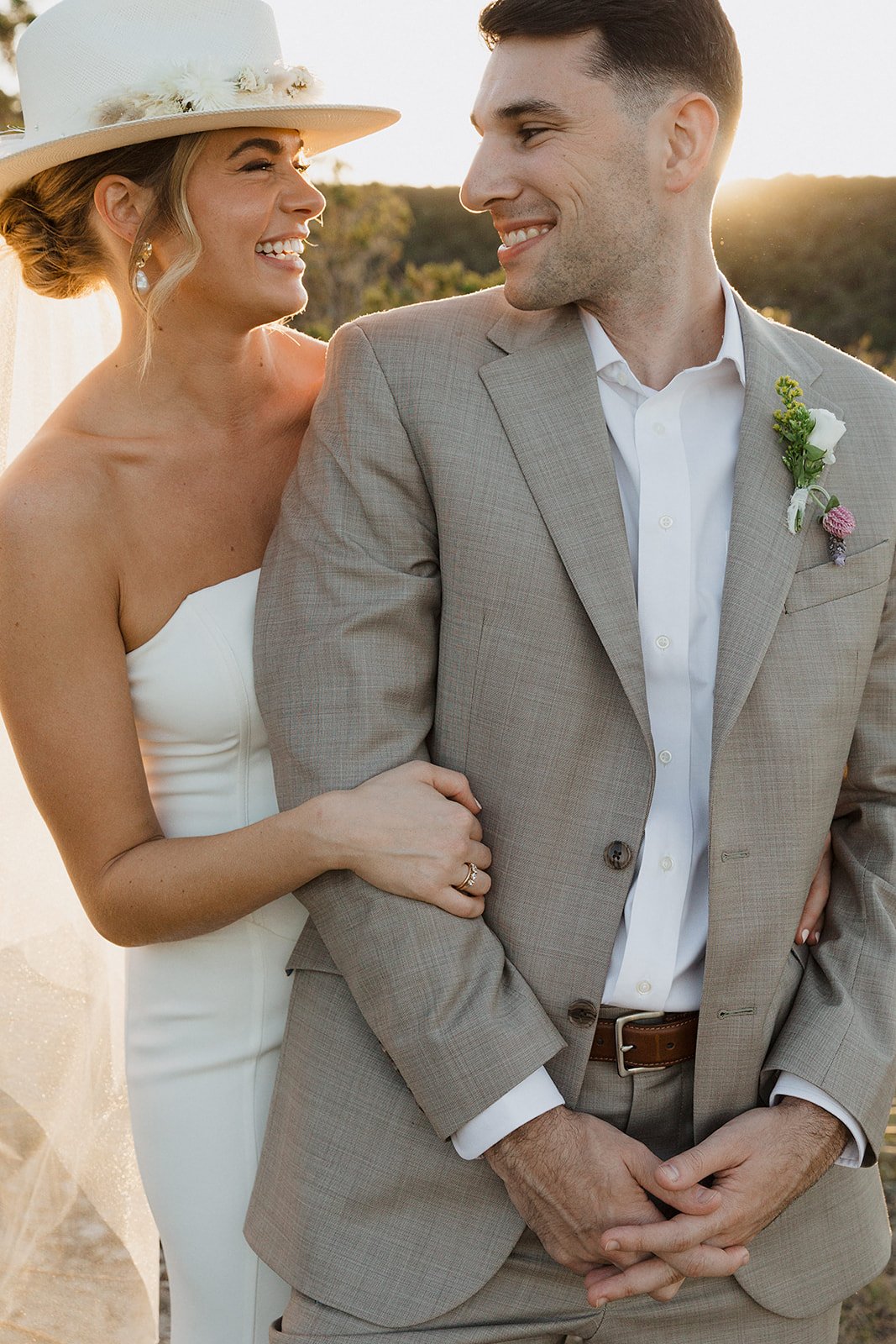 A bride and groom during their wedding, outdoors at sunset, with the bride smiling at the groom, both dressed in wedding attire.