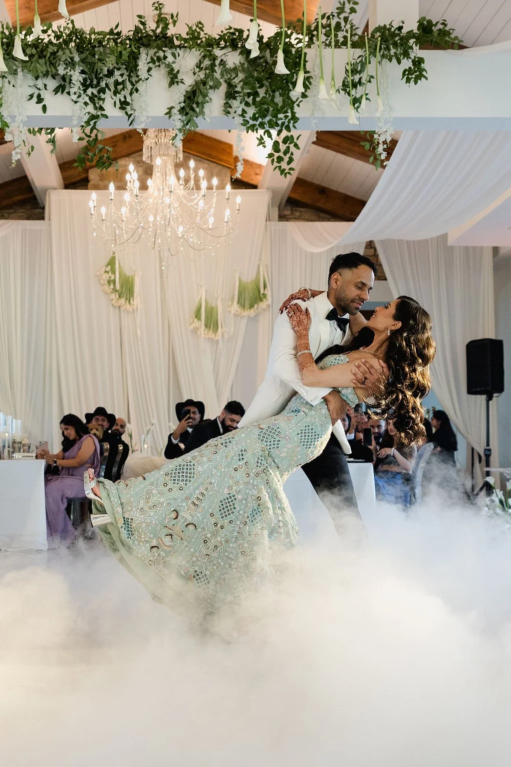 A wedding couple dancing in a decorated venue with a chandelier and white drapery, surrounded by guests and floral decorations.