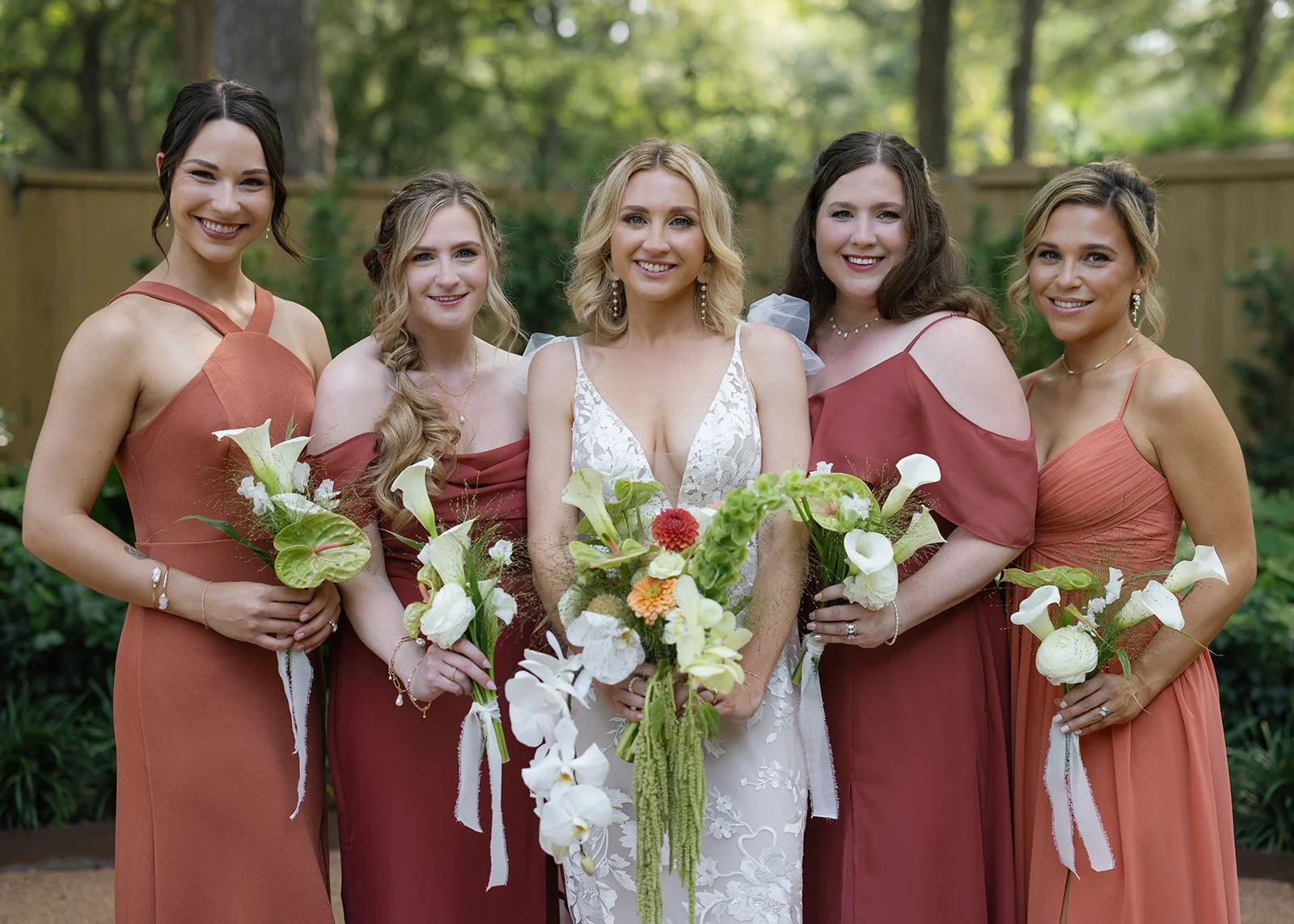 A bride in a white lace wedding dress standing with five bridesmaids in rust-colored dresses, all holding bouquets of flowers, outdoors in a garden.