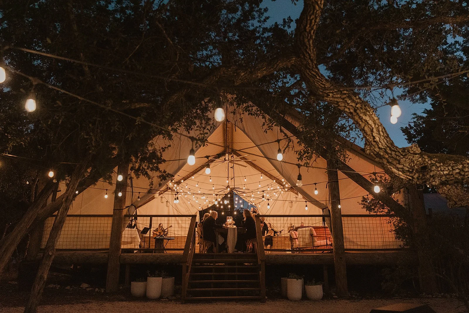 A nighttime outdoor event inside a large white tent adorned with string lights. Guests are seated at a table, while musicians perform on a small stage, with some playing guitars. The tent is surrounded by trees and potted plants, and a staircase lead