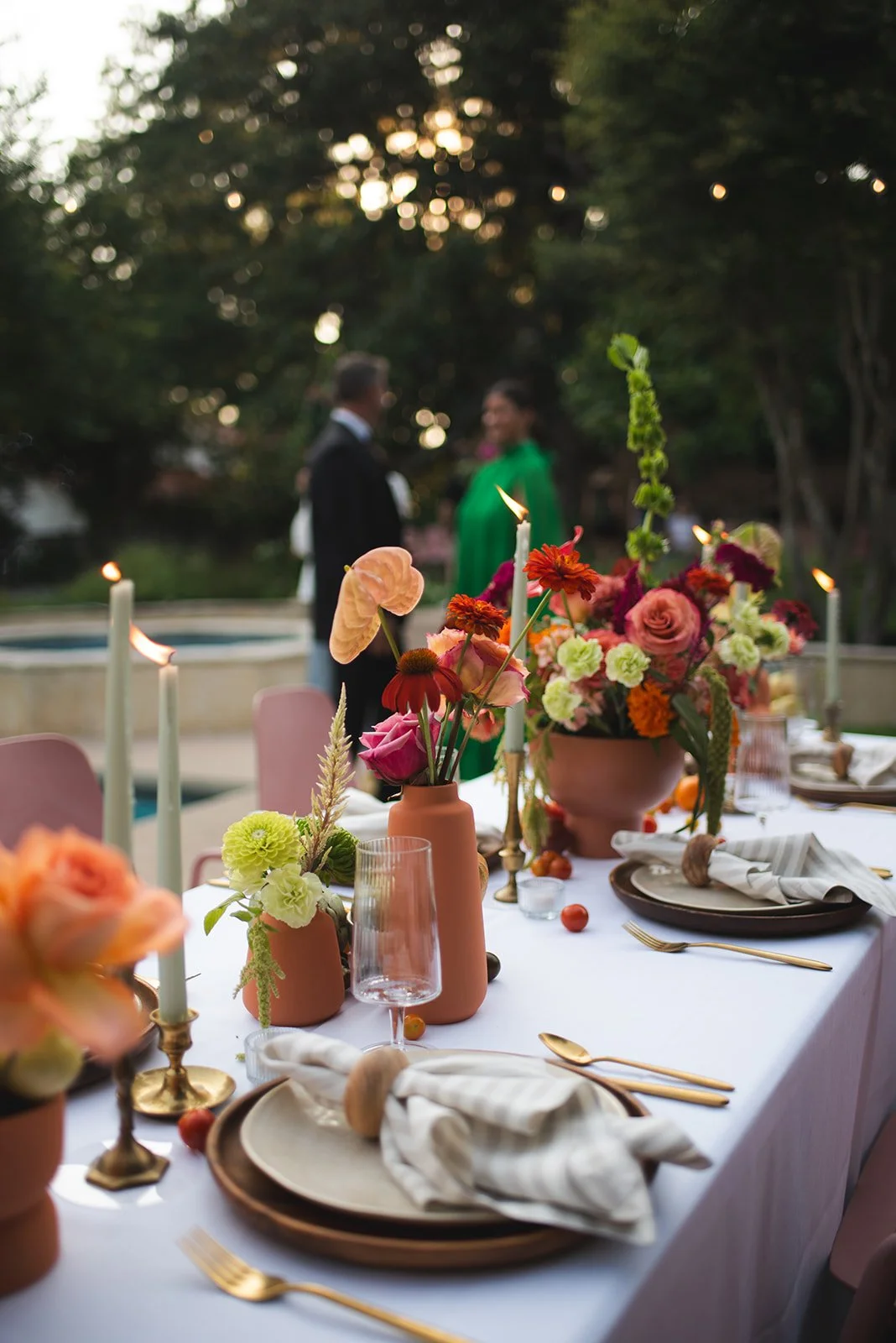 Elegant outdoor dinner table with pink, orange, and green floral arrangements, candles, gold flatware, and white napkins, with blurred guests talking in the background under sunset lighting.