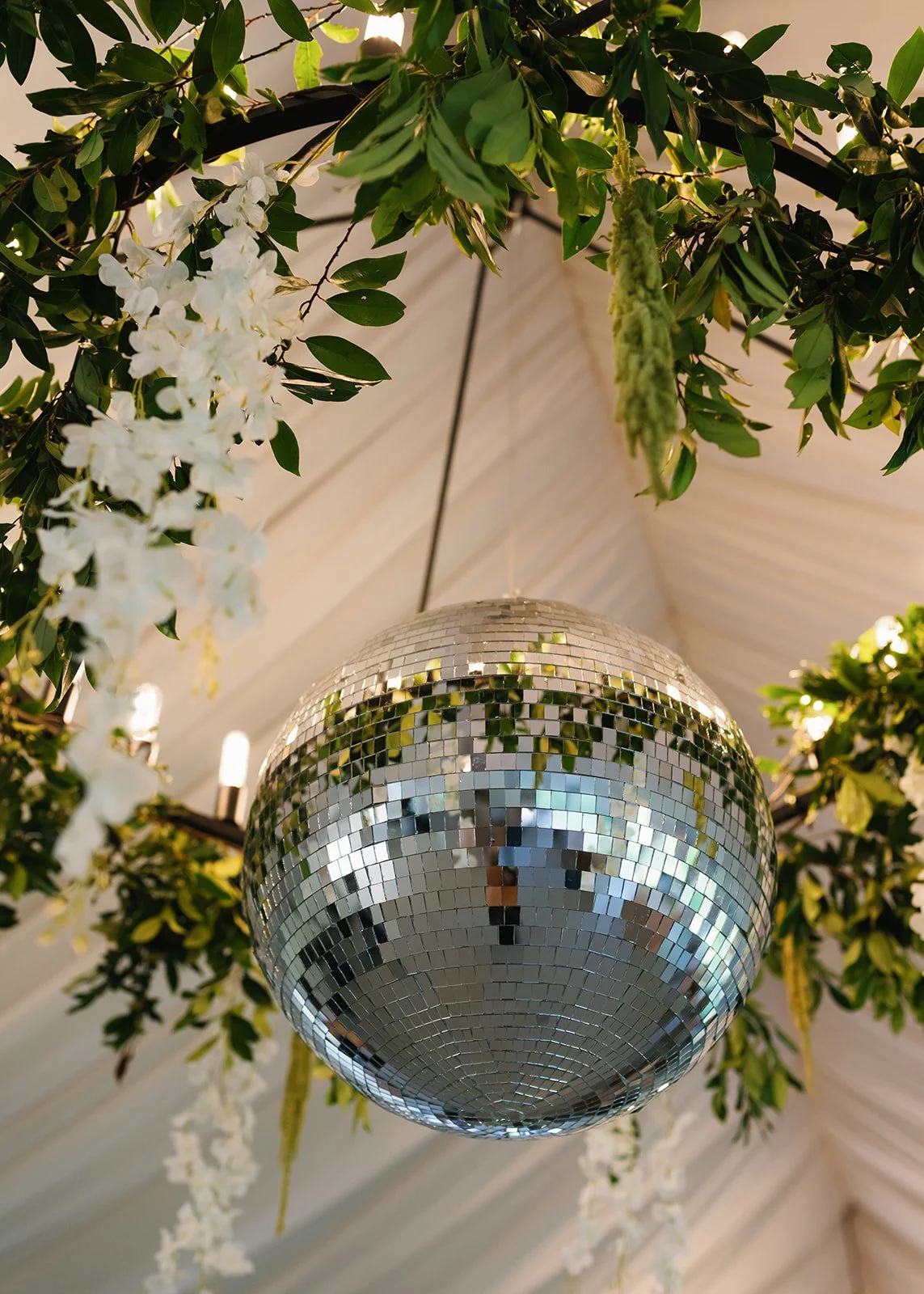 A silver disco ball hanging from the ceiling among green leaves and white flowers in a decorated indoor setting.