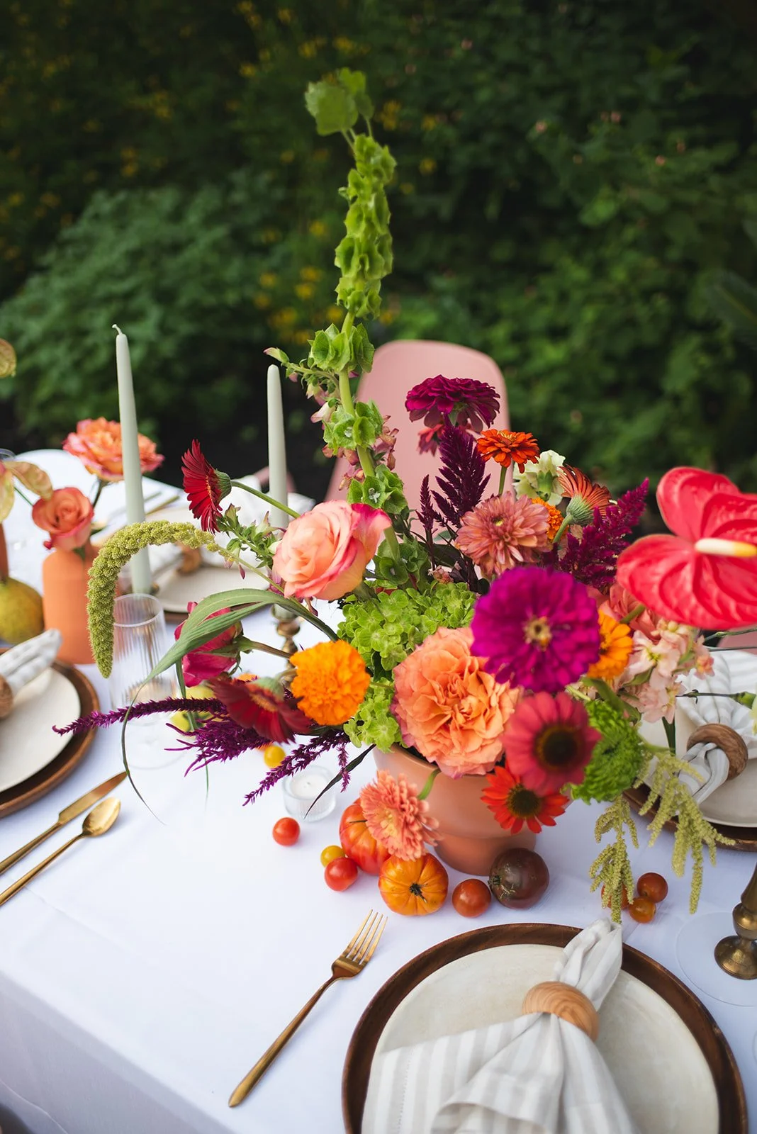 A table decorated with a large colorful flower arrangement in a pink vase, surrounded by small tomatoes, including orange and red varieties, with table settings that include gold utensils, white napkins, and plates, on a white tablecloth outdoors ami