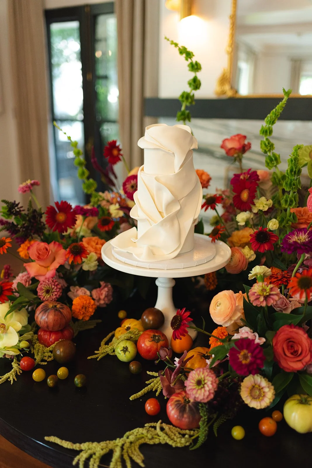 A white wedding cake with elegant ruffled design on a white cake stand, surrounded by colorful flowers and assorted small vegetables and fruits on a black table.