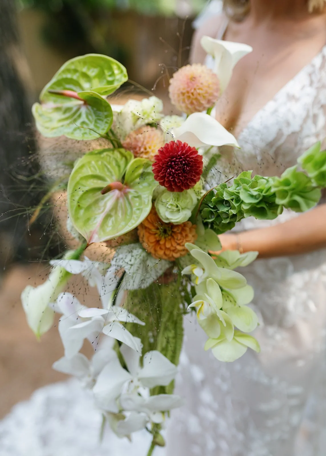 Bride holding a cascading bouquet of white calla lilies, orange dahlias, green anthuriums, pink and peach dahlias, and greenery, wearing a lace wedding dress.