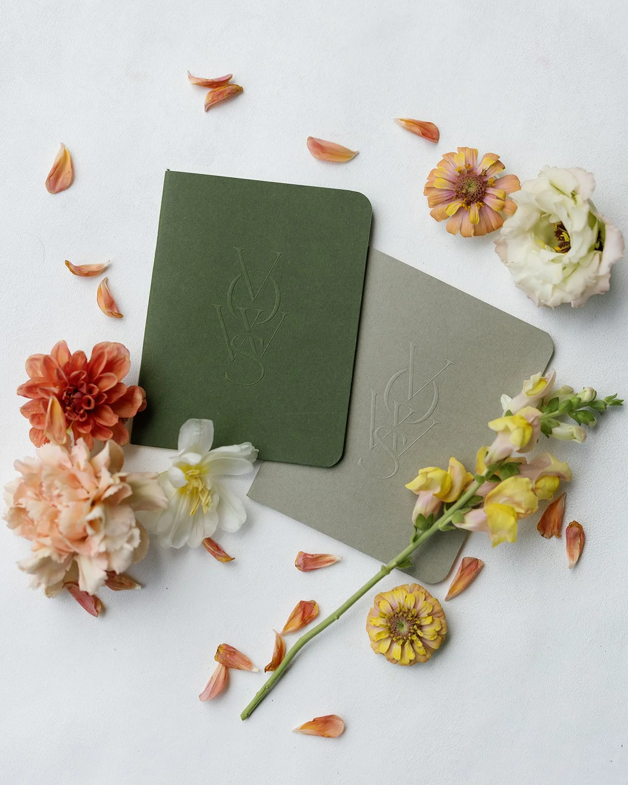 Two folded stationery cards, one dark green and one light gray, surrounded by assorted flowers and scattered flower petals on a white background.