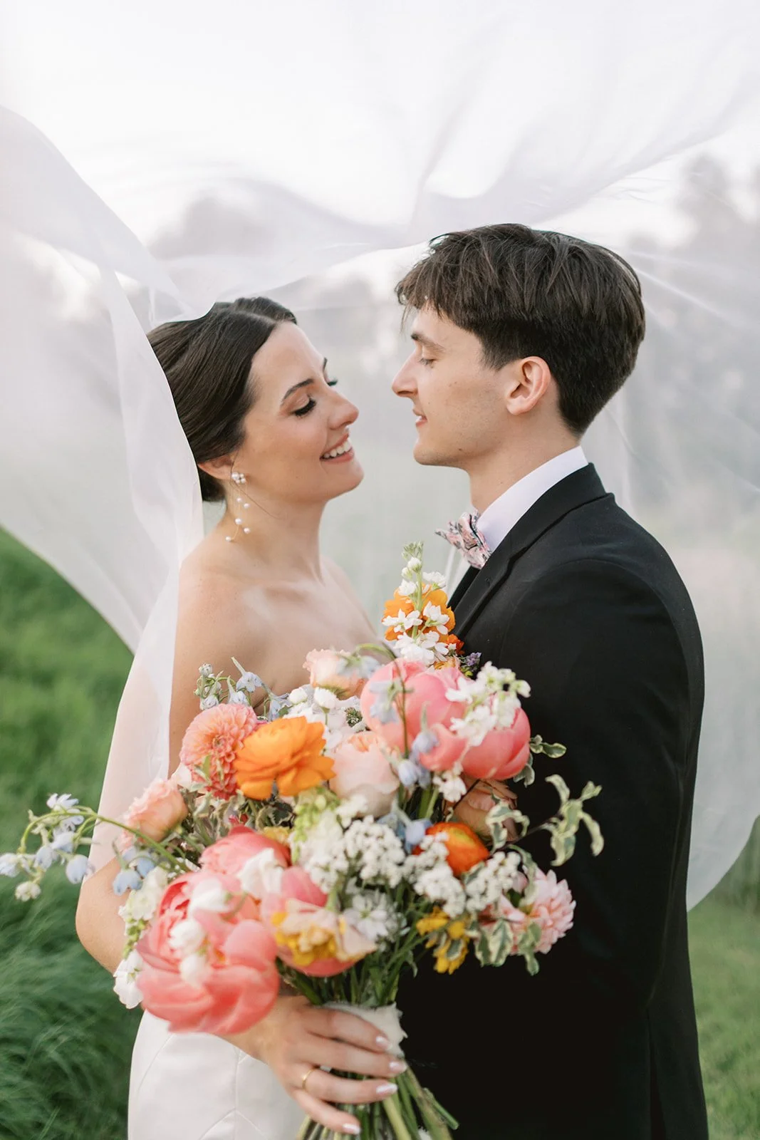 A bride and groom face each other, smiling, with a flowing white veil around them, outdoors with green grass in the background. The bride holds a large colorful bouquet of flowers.